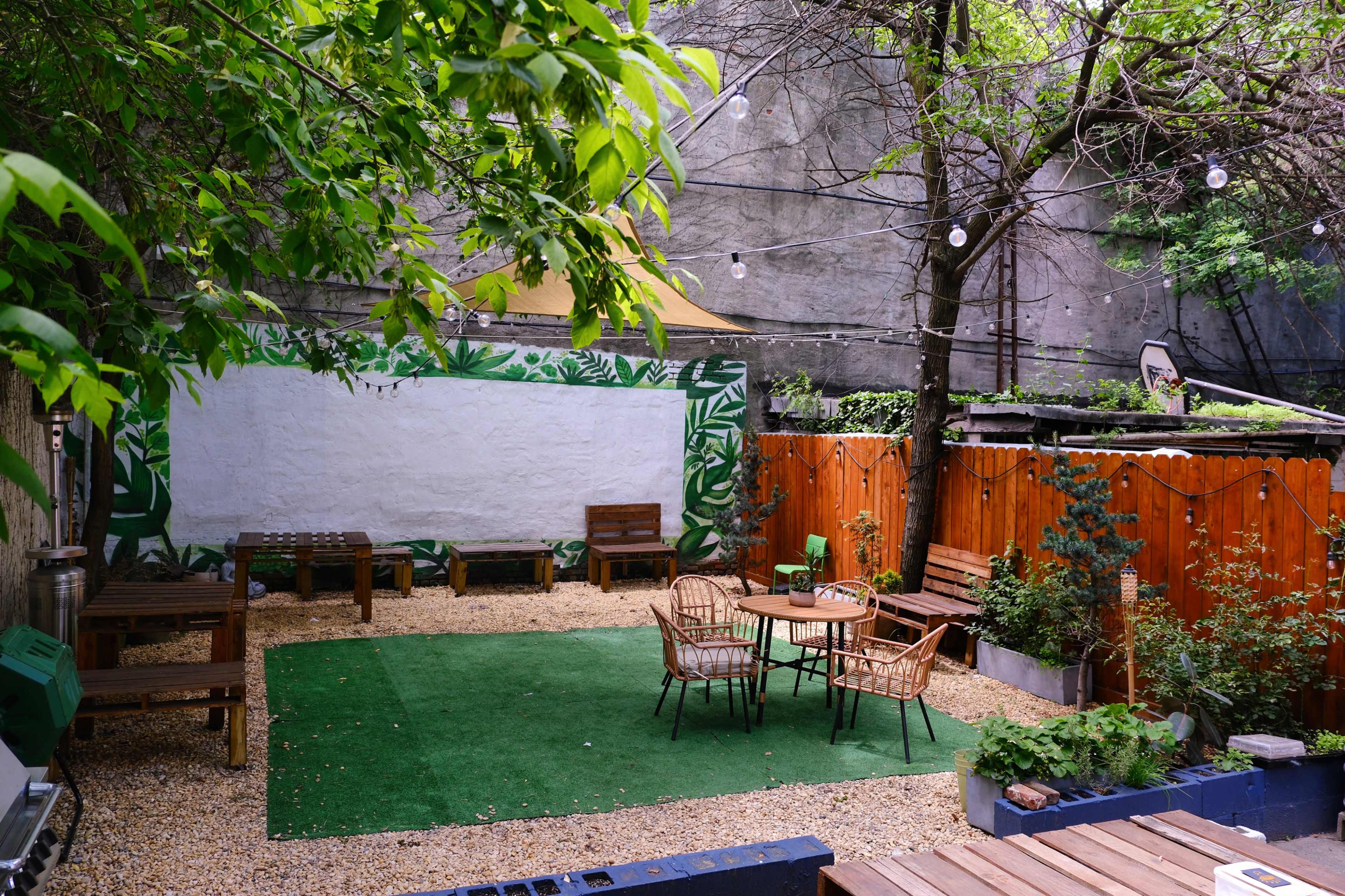 A small outdoor space featuring green grass, wooden seating, potted plants, and a mural on a wall surrounded by trees and string lights.
