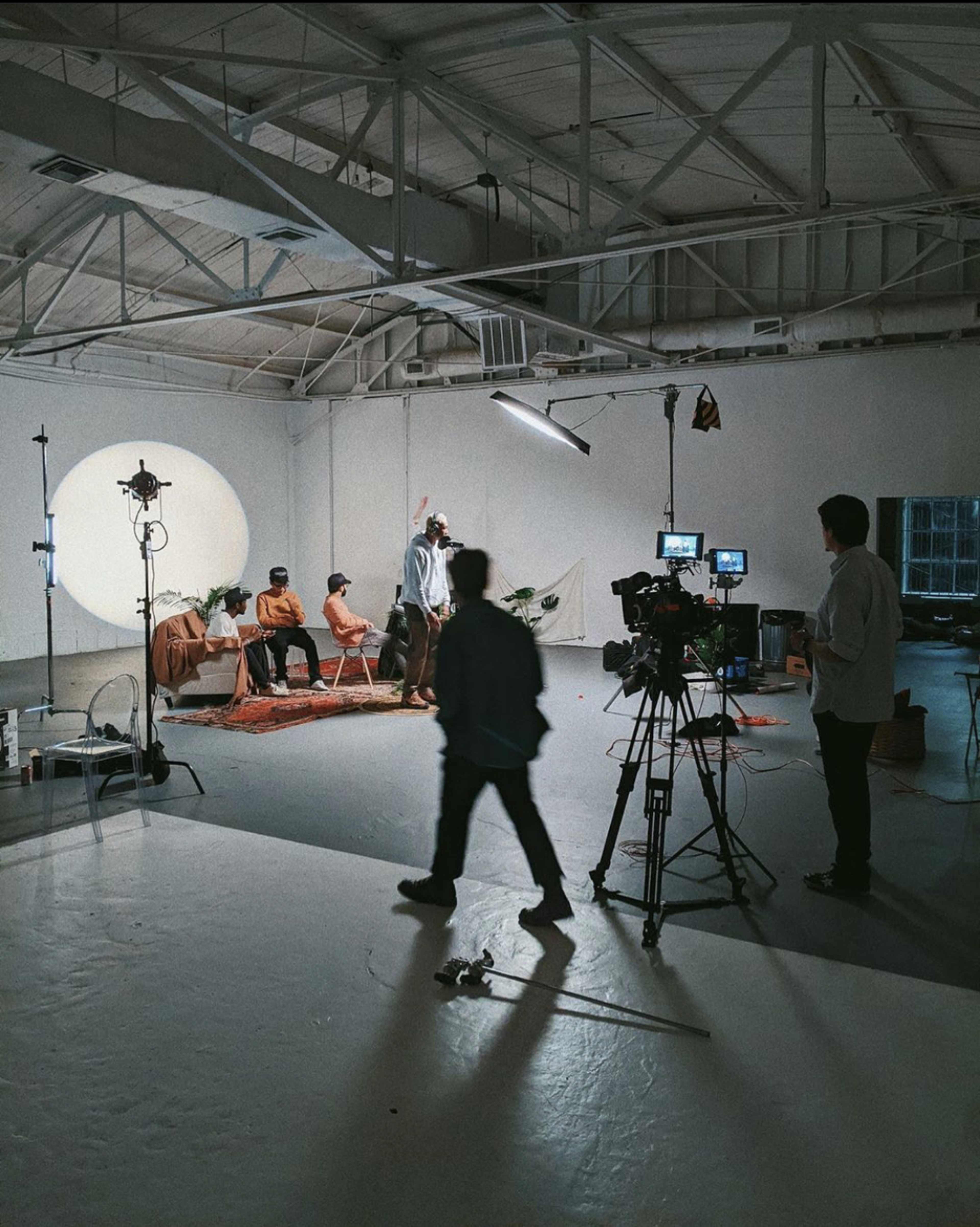 A film crew captures a scene with a group of four individuals sitting on chairs in a minimalist studio with a circular backdrop and various cameras set up.