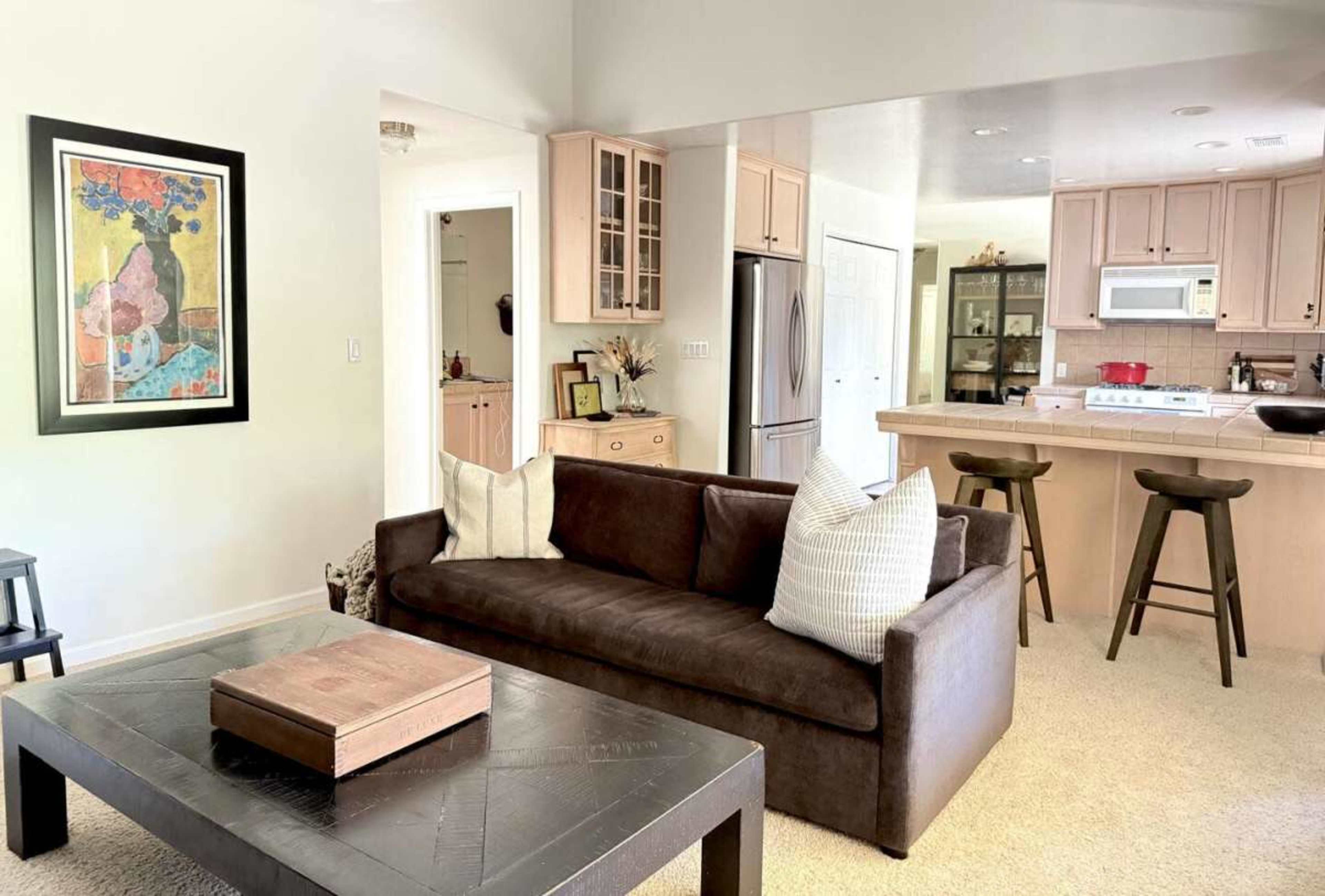 A brown sofa with white pillows sits in a cozy living area, facing a black coffee table, with an open kitchen and dining space visible in the backdrop.