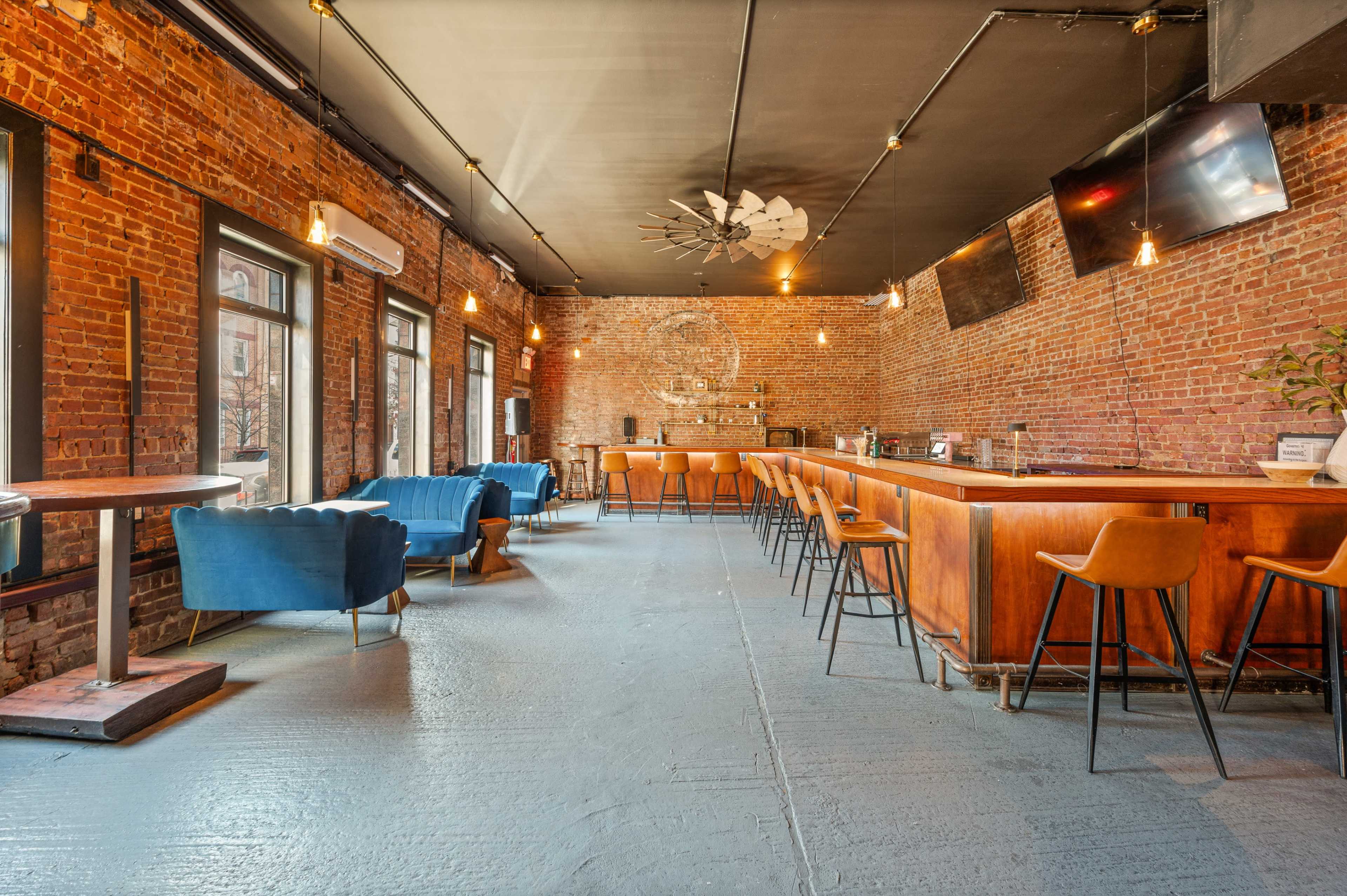 The image shows a spacious bar area with exposed brick walls, wooden furniture, and stools arranged around a central bar counter.
