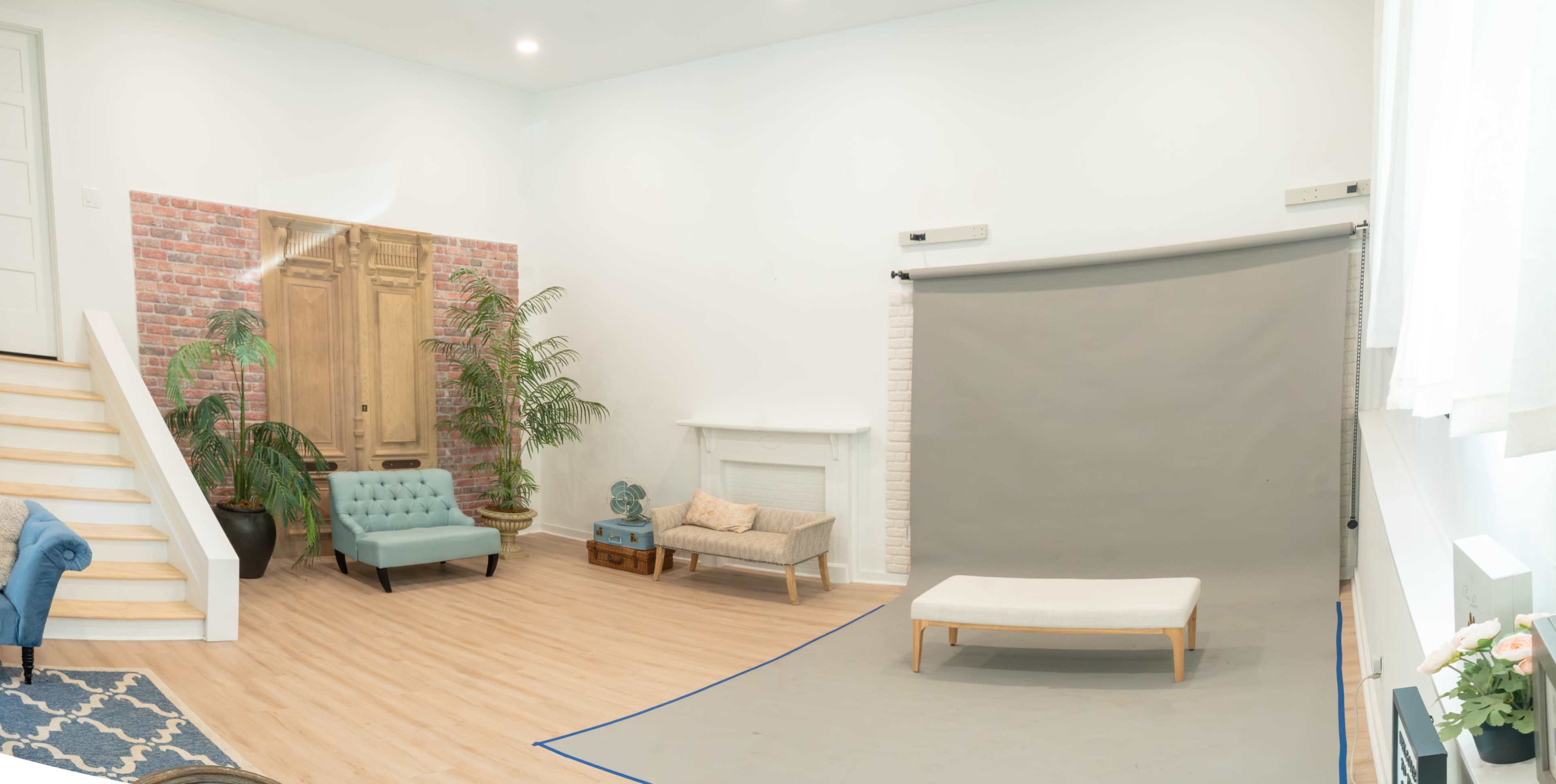 The image shows a modern studio space featuring light wood flooring, a gray backdrop, a sofa, an armchair, and decorative elements like plants and a brick wall.