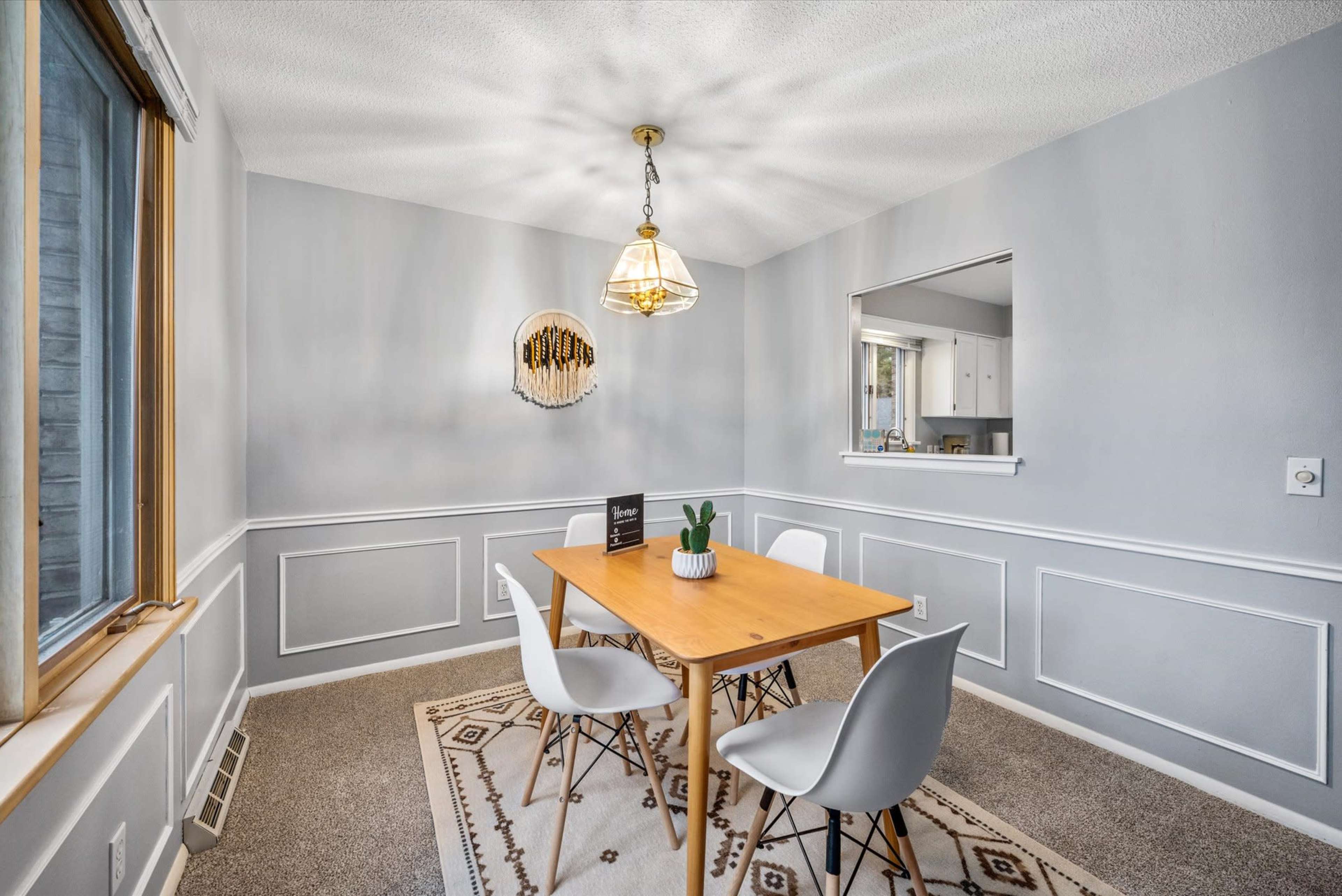 A dining area features a wooden table surrounded by white chairs, with a decorative light fixture overhead and a window providing natural light.
