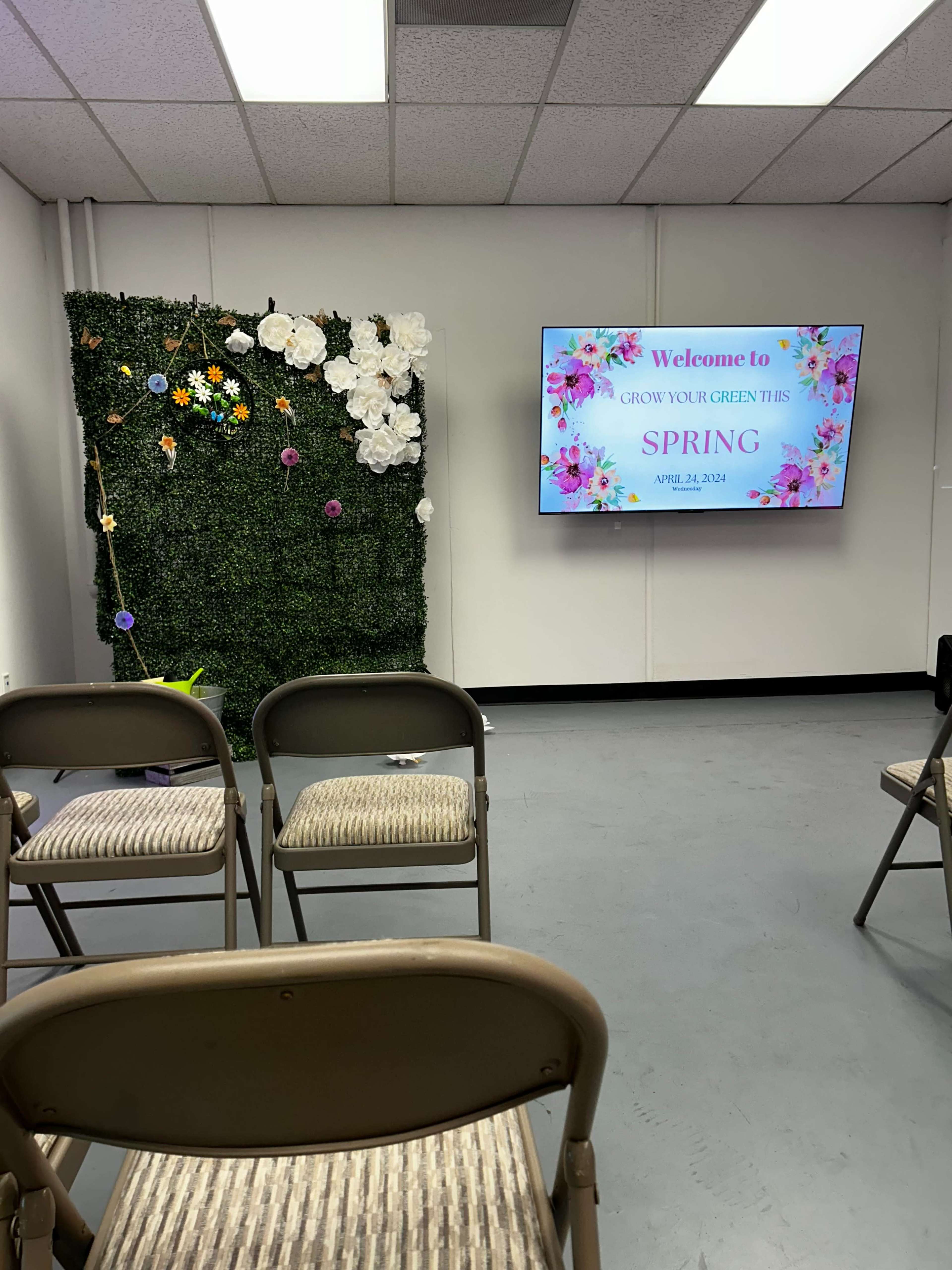 A room setup for an event with a decorative green wall adorned with flowers, a large screen displaying "Welcome to Grow Your Green This Spring," and several empty chairs arranged in front.