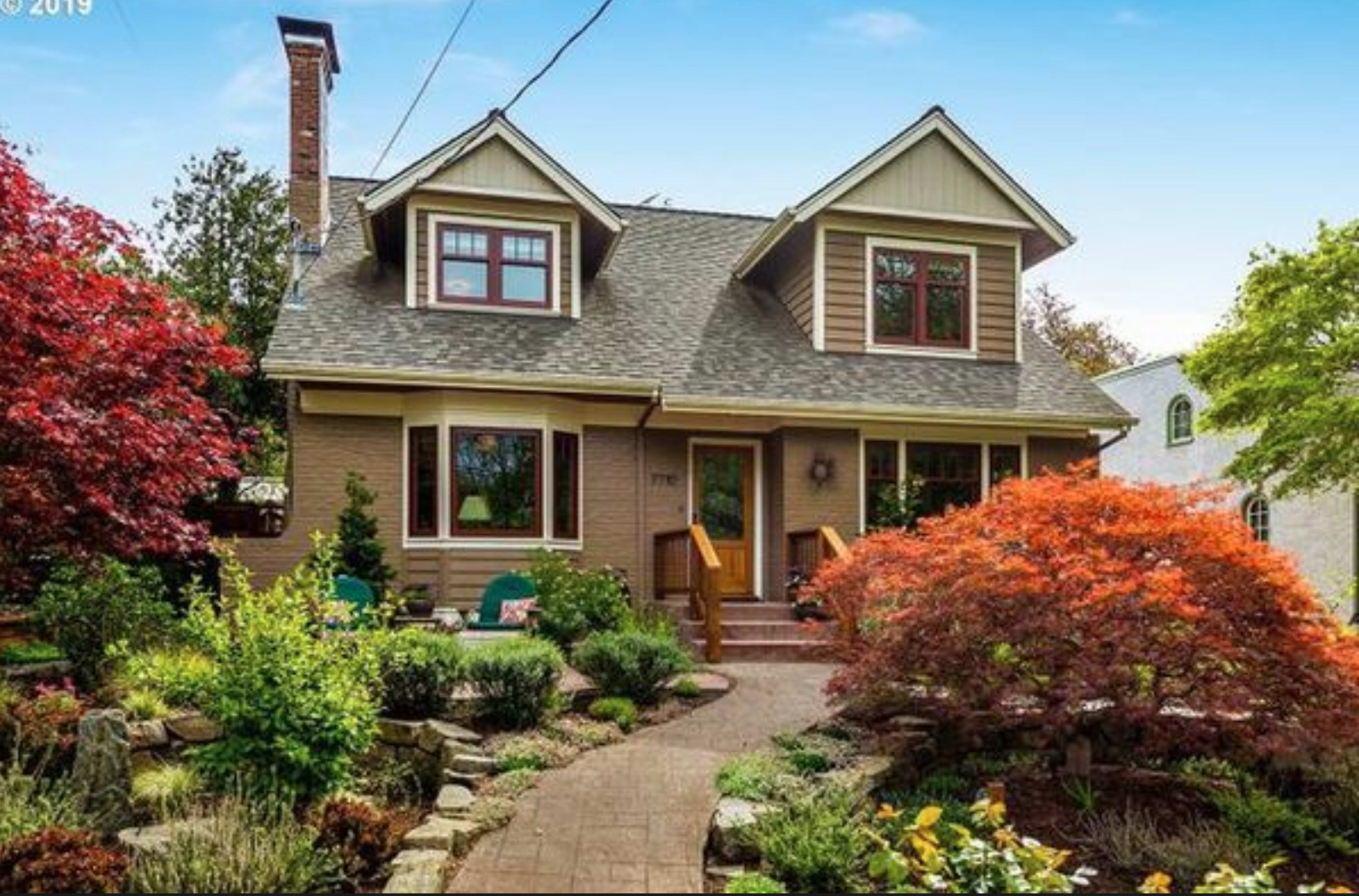The image shows a two-story house with a sloped roof, surrounded by a landscaped garden featuring colorful shrubs and a stone pathway.
