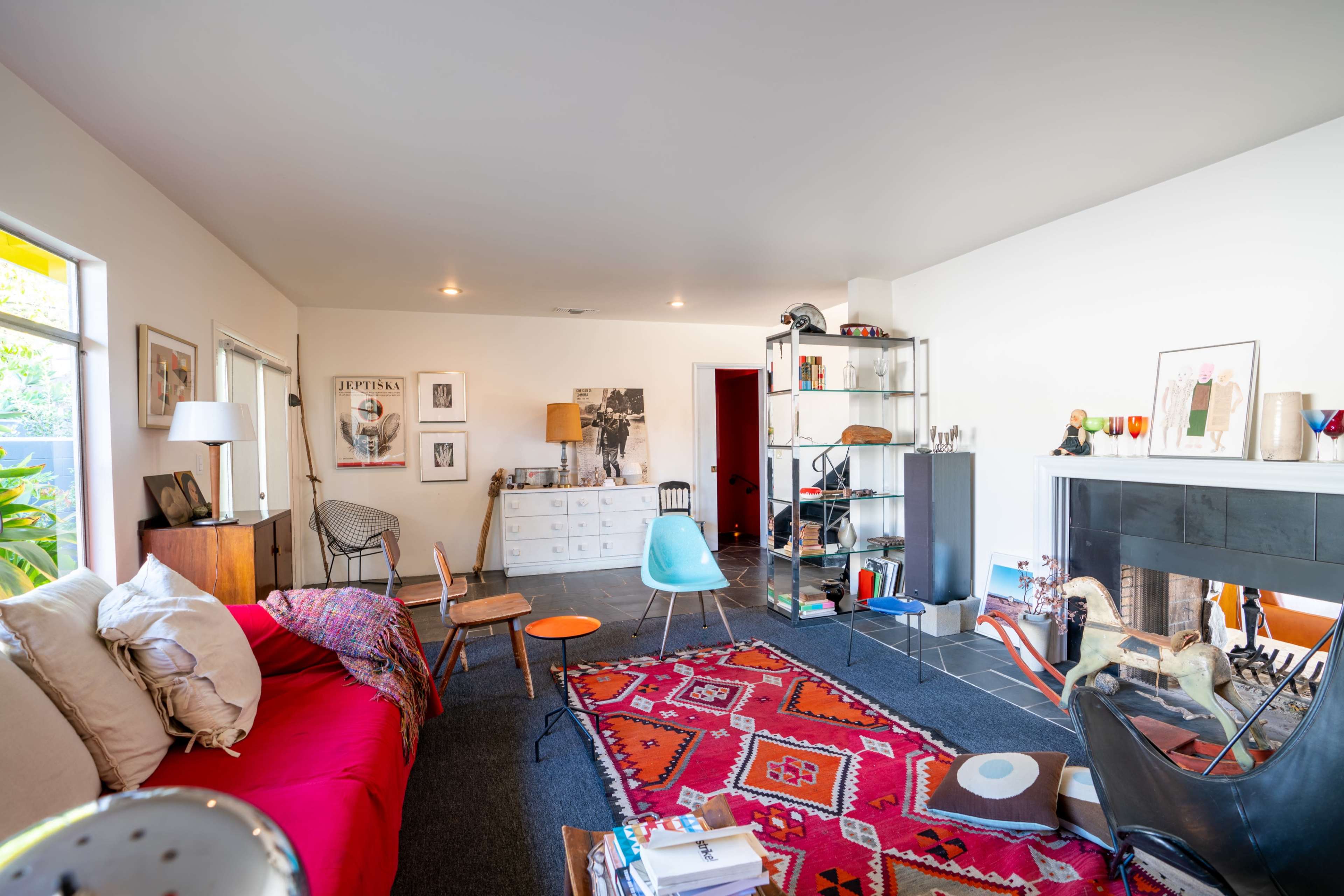 A spacious living room featuring a red sofa, a patterned rug, various chairs, and shelves filled with decorations and artwork.
