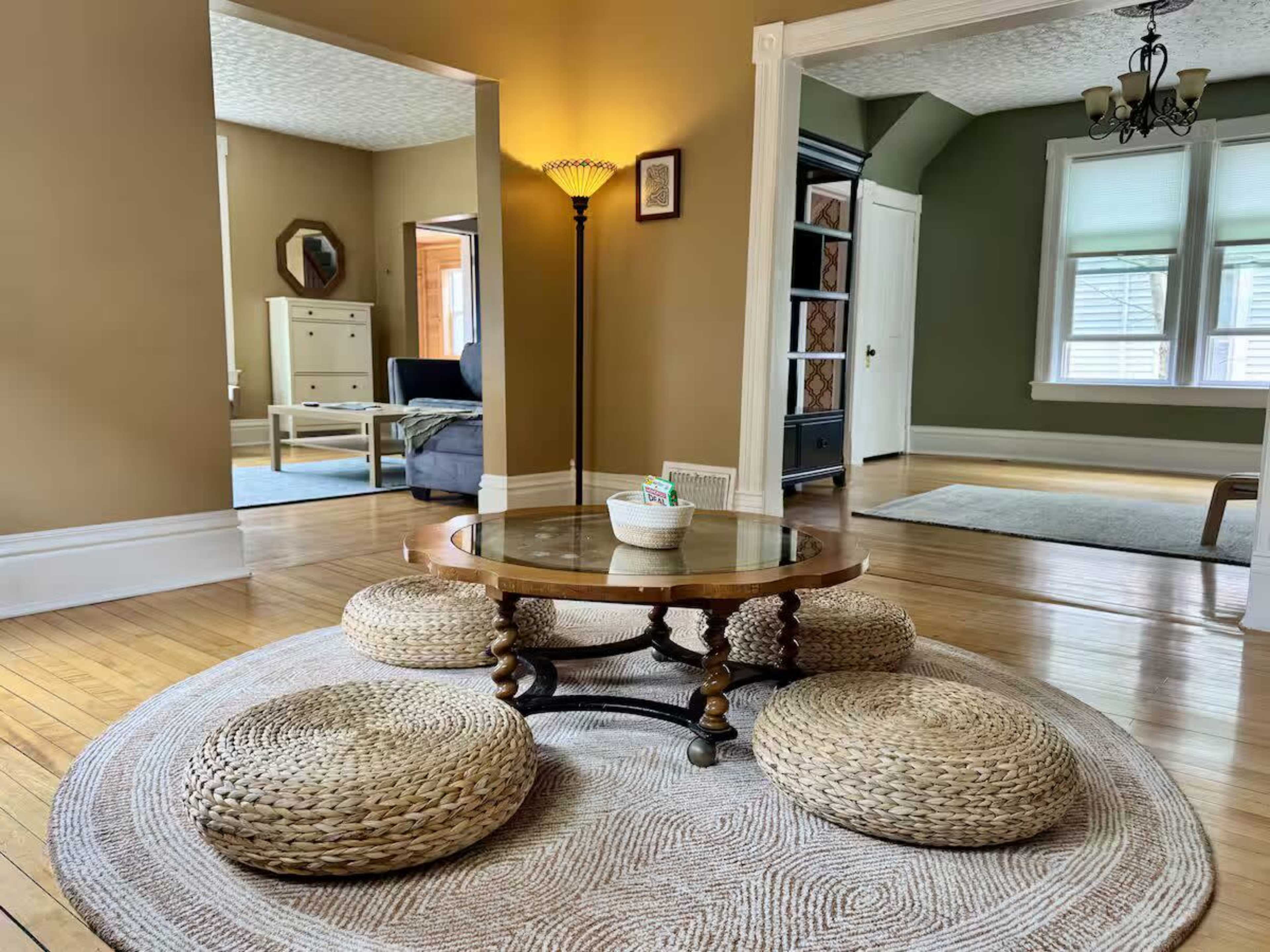 The image shows a living space with a round table surrounded by woven seating on a circular rug, with light walls and a doorway leading to another room.