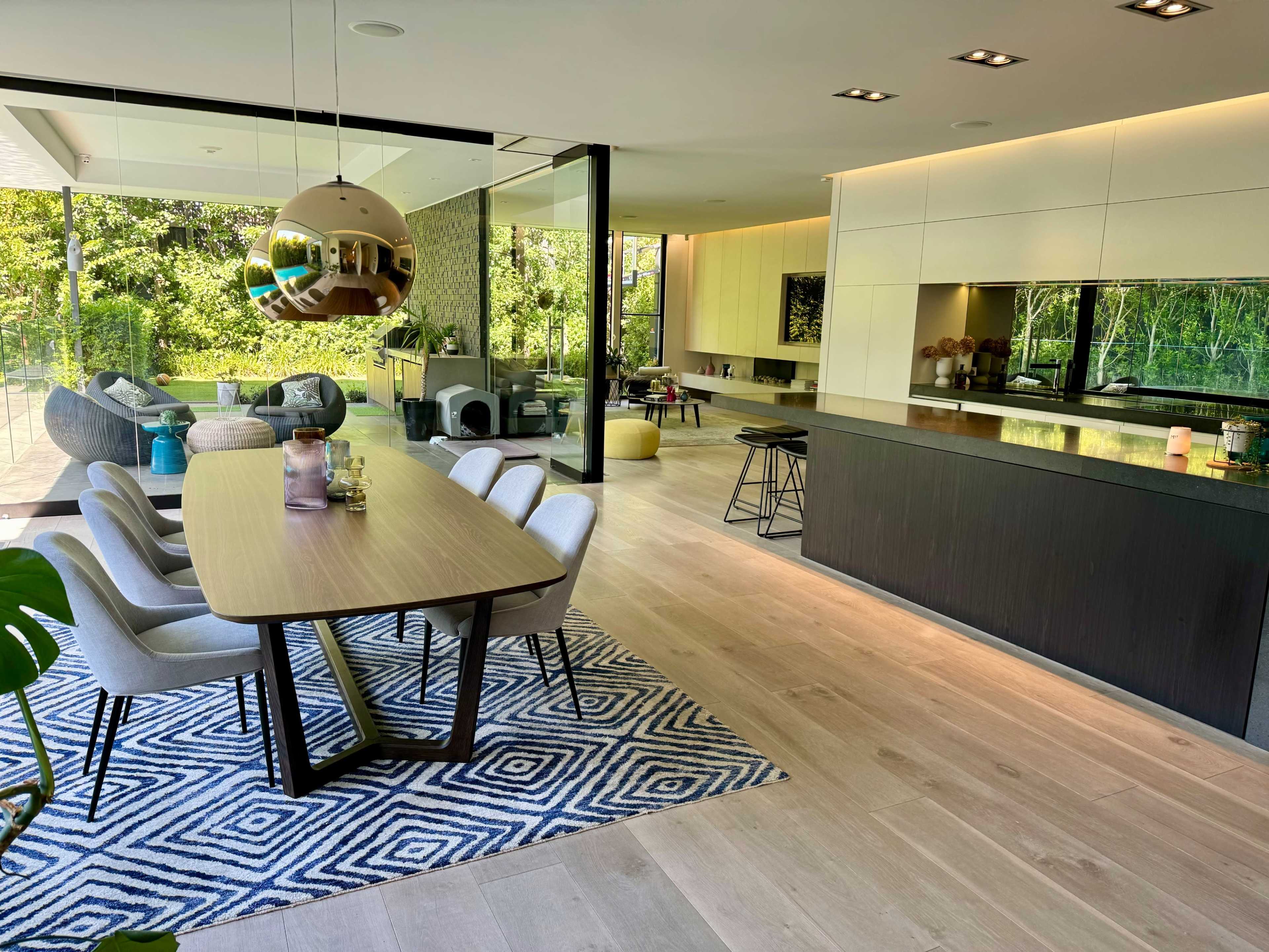 A modern dining area with a large table, stylish chairs, and an open kitchen overlooking a garden through large glass windows.