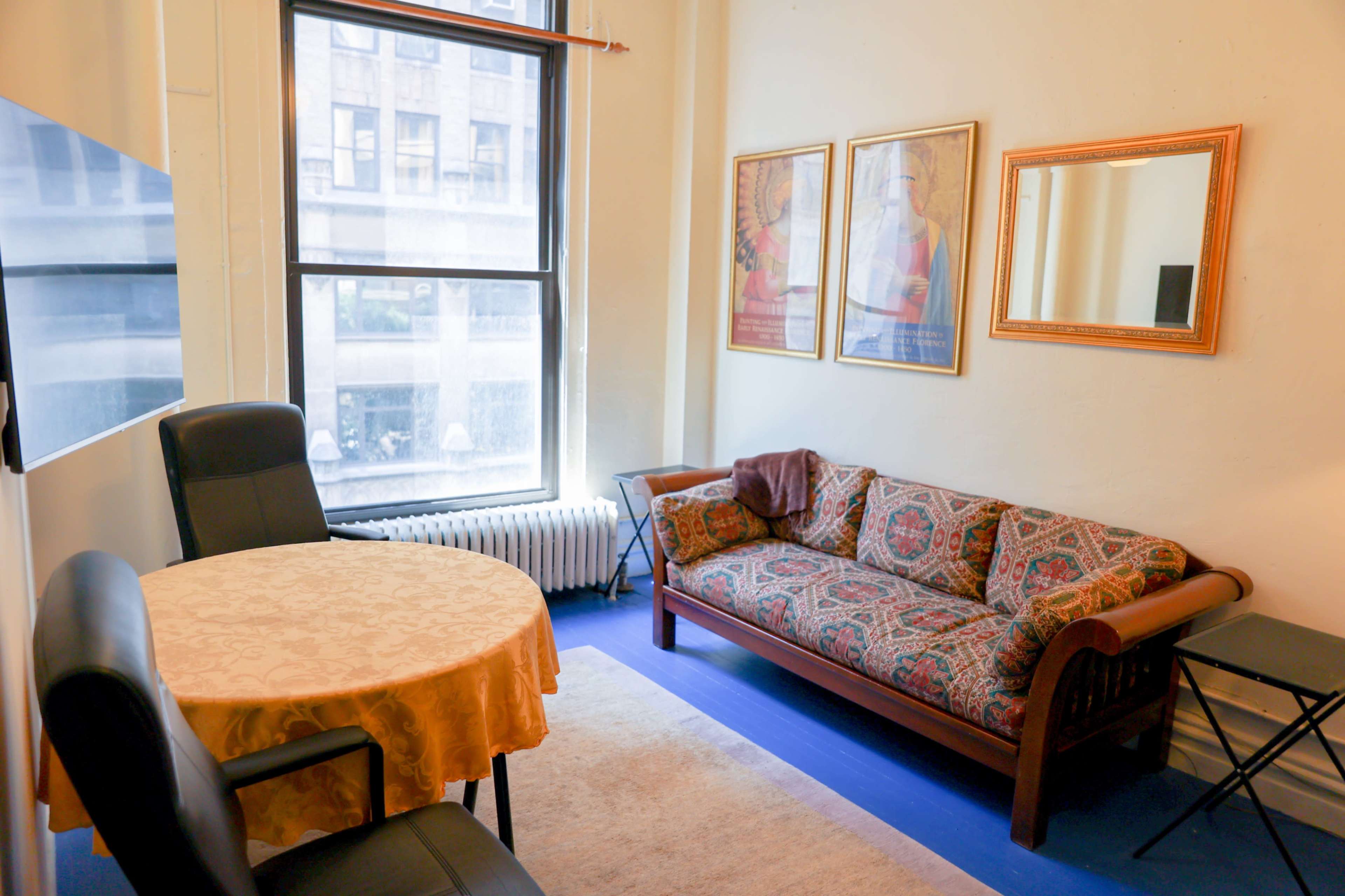 The image shows a small room with a patterned couch, a round dining table, and framed art on the walls, along with a large window letting in natural light.