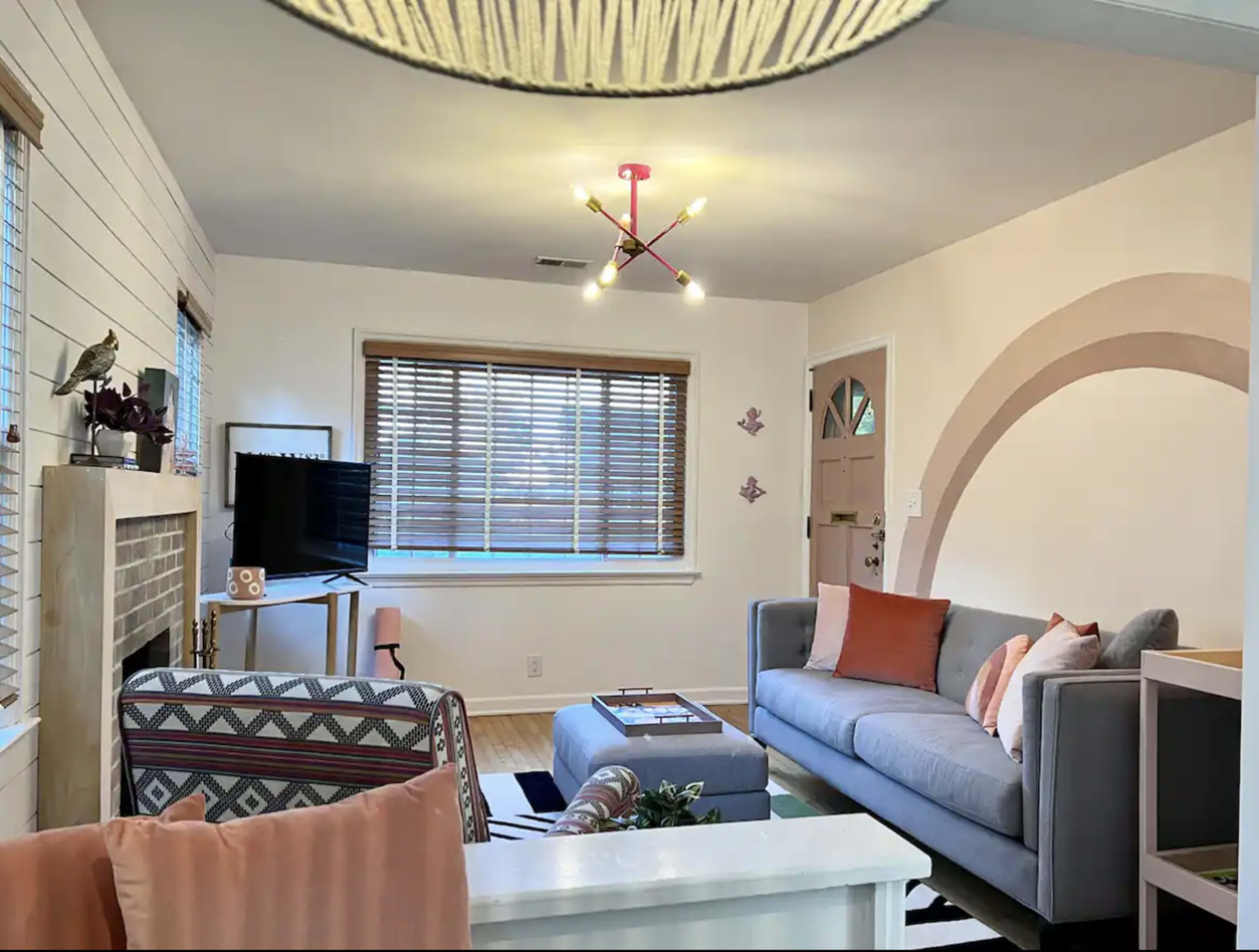 A living room with a gray sofa, a patterned armchair, a television on a stand, and a coffee table, all set against a backdrop of light-colored walls and wooden blinds.