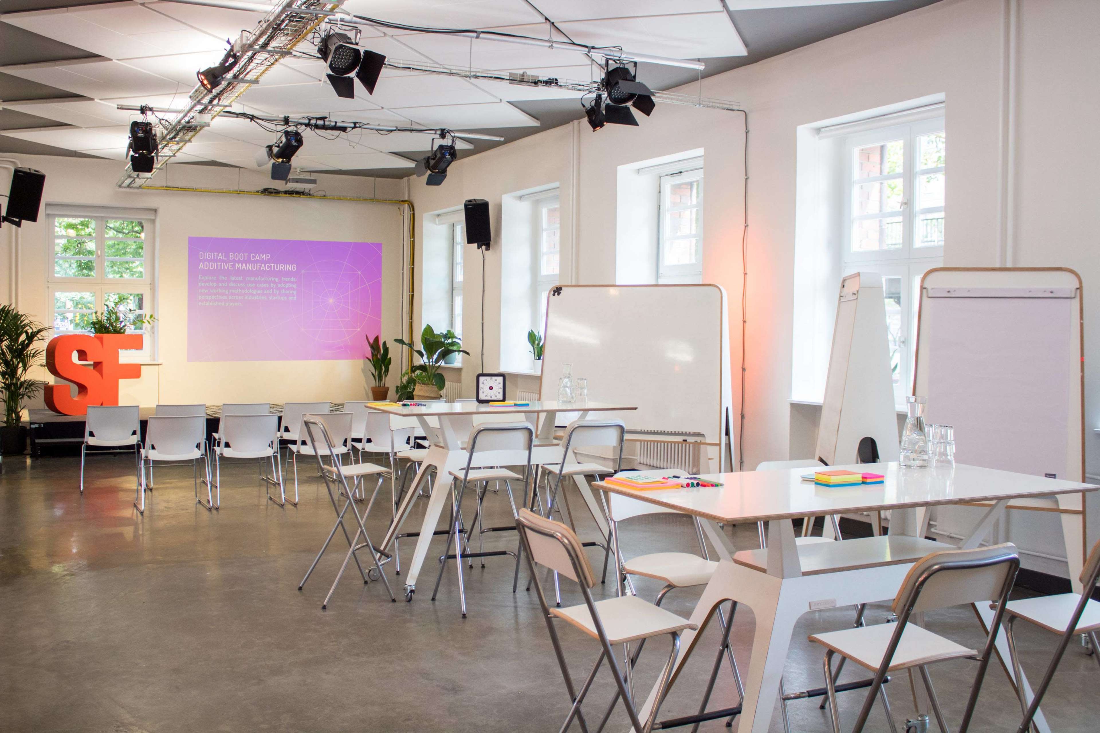 A modern, well-lit seminar room set up for a presentation, featuring chairs, tables, and a large screen displaying a digital presentation.