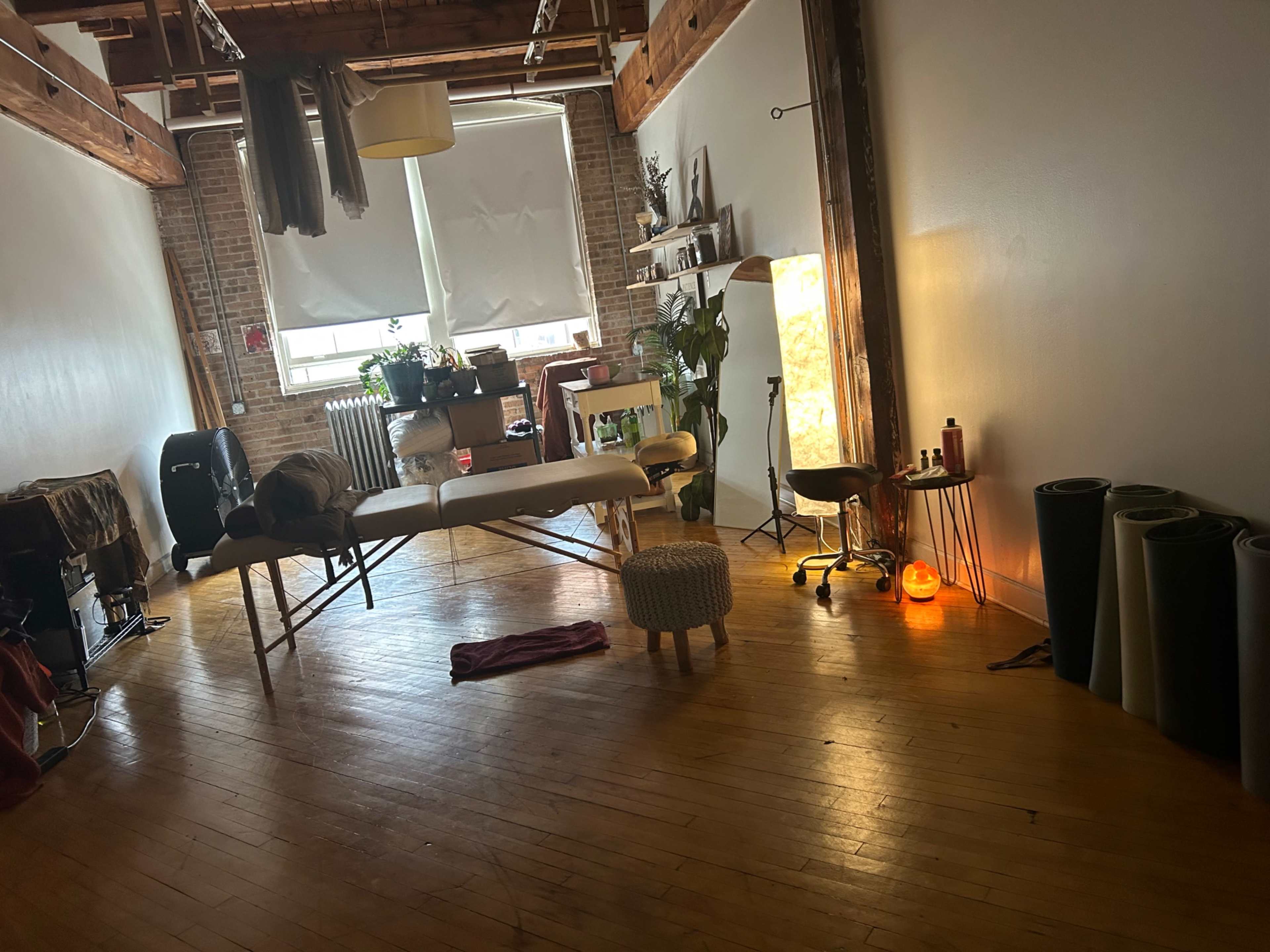 The image shows a spacious interior room with wooden floors, featuring a massage table, a small stool, and yoga mats rolled up against the wall.