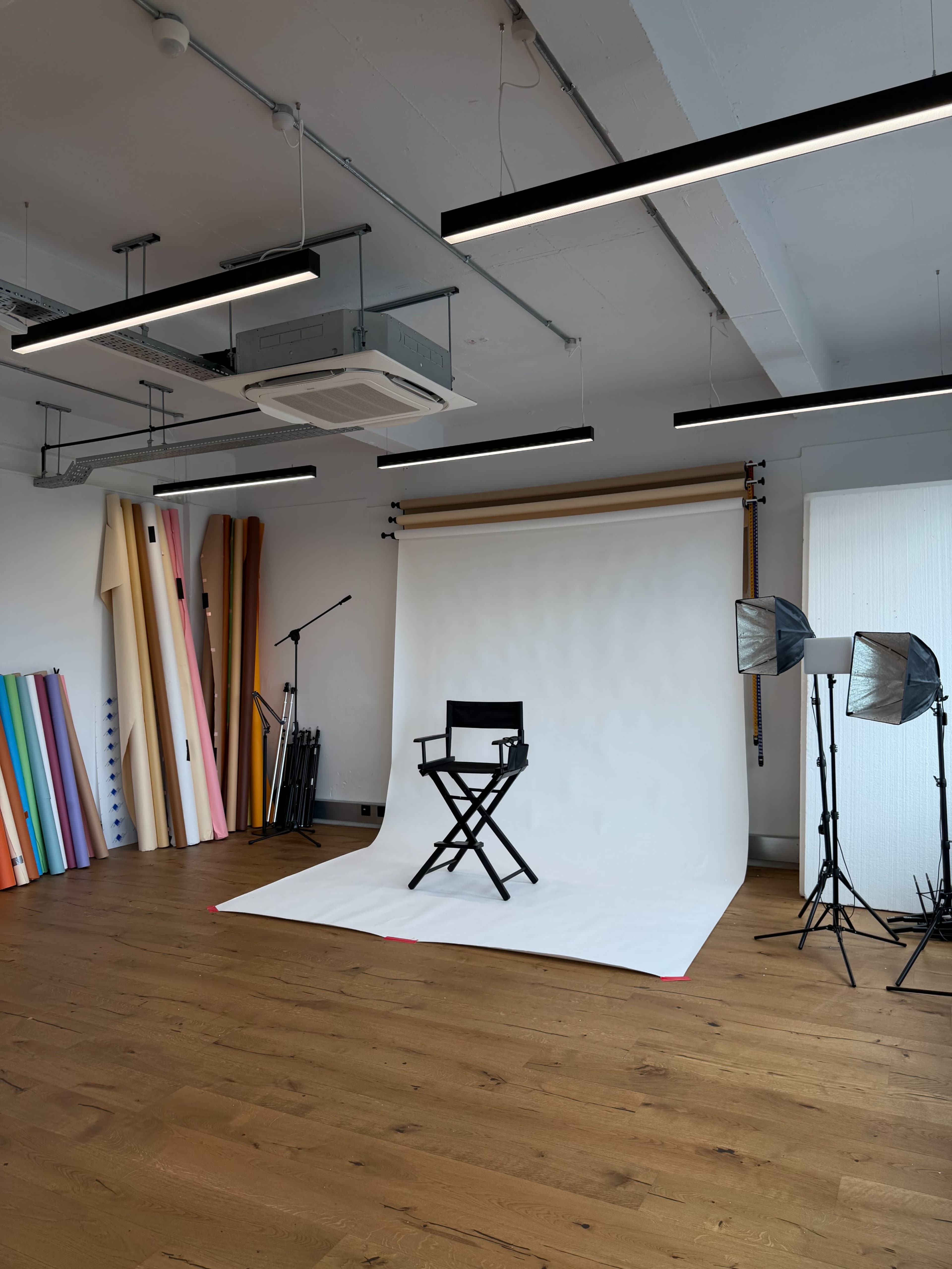 The image shows a photography studio equipped with a director's chair, backdrop, lighting equipment, and rolls of colored paper against the wall.