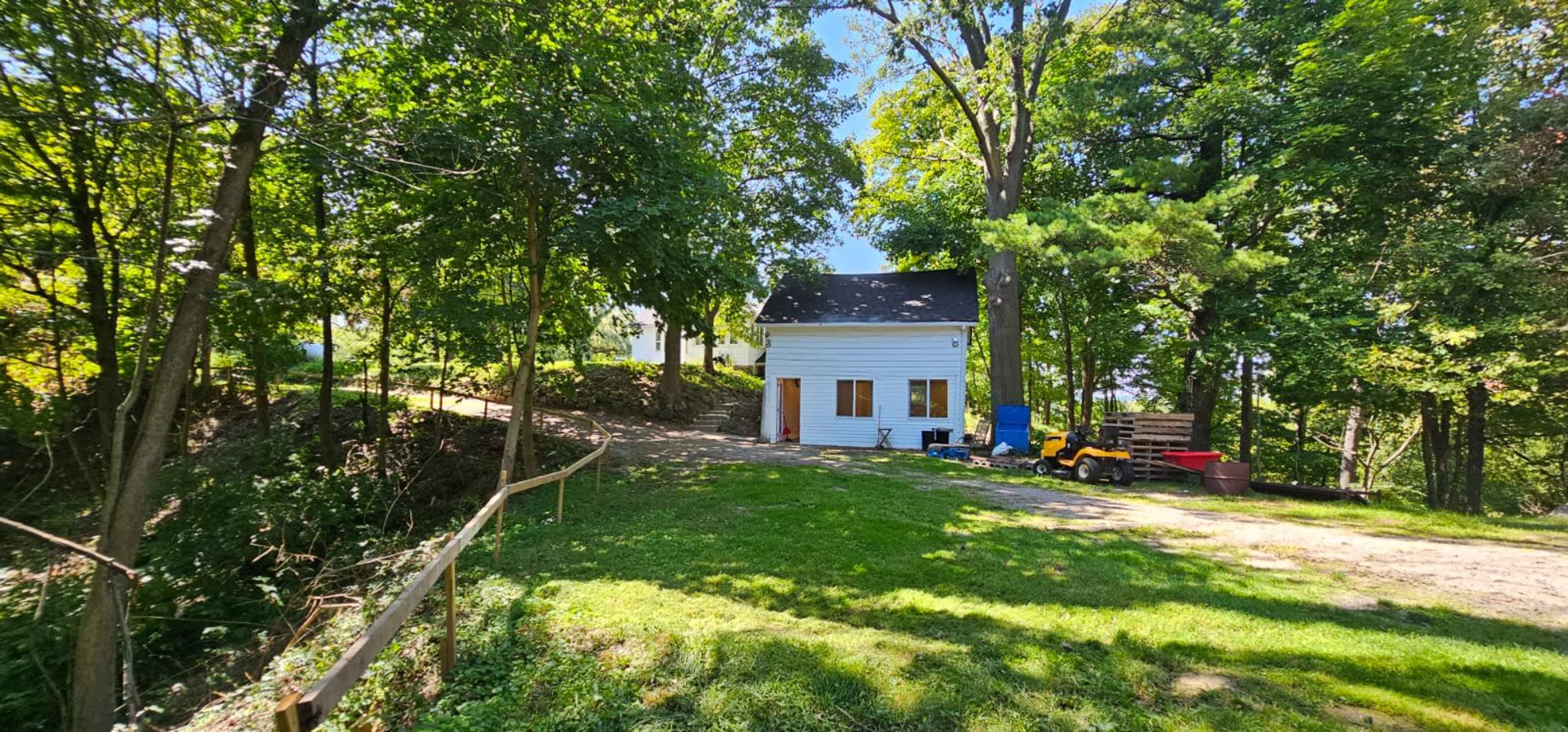 A small white house is situated among trees on a grassy area, with a gravel path leading up to it.