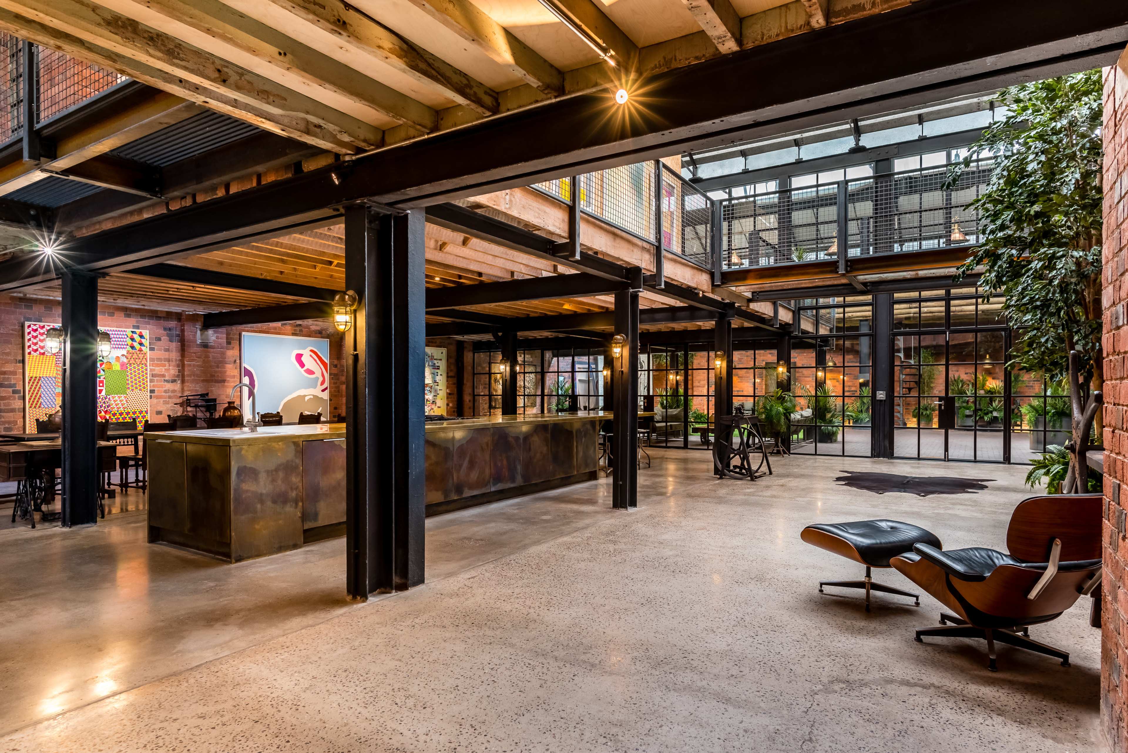 A spacious industrial-style interior with exposed brick walls, large windows, and a mix of modern furniture and plants.