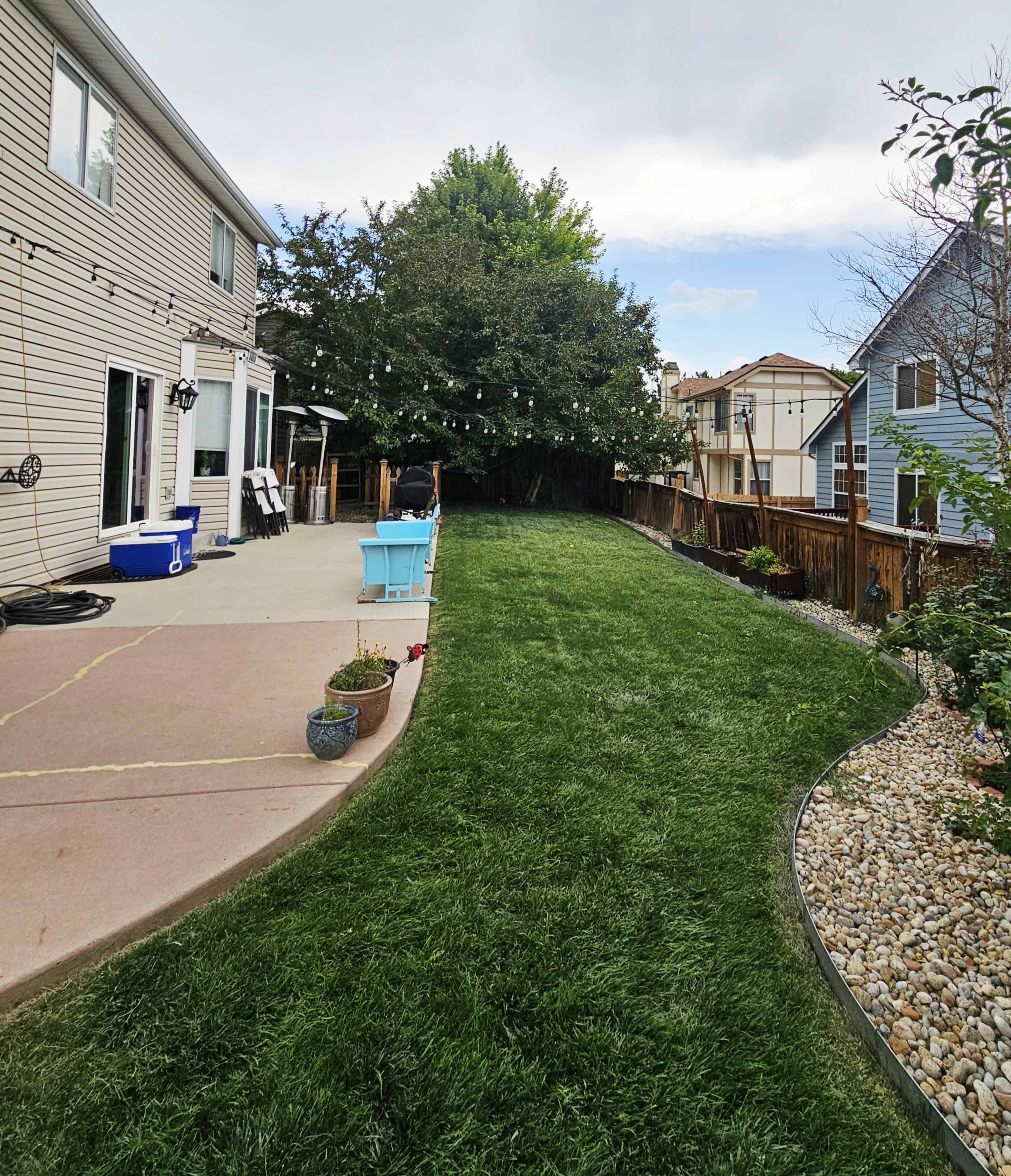 The image shows a landscaped backyard with a green lawn, a patio area adjacent to a house, and decorative stones along the border.