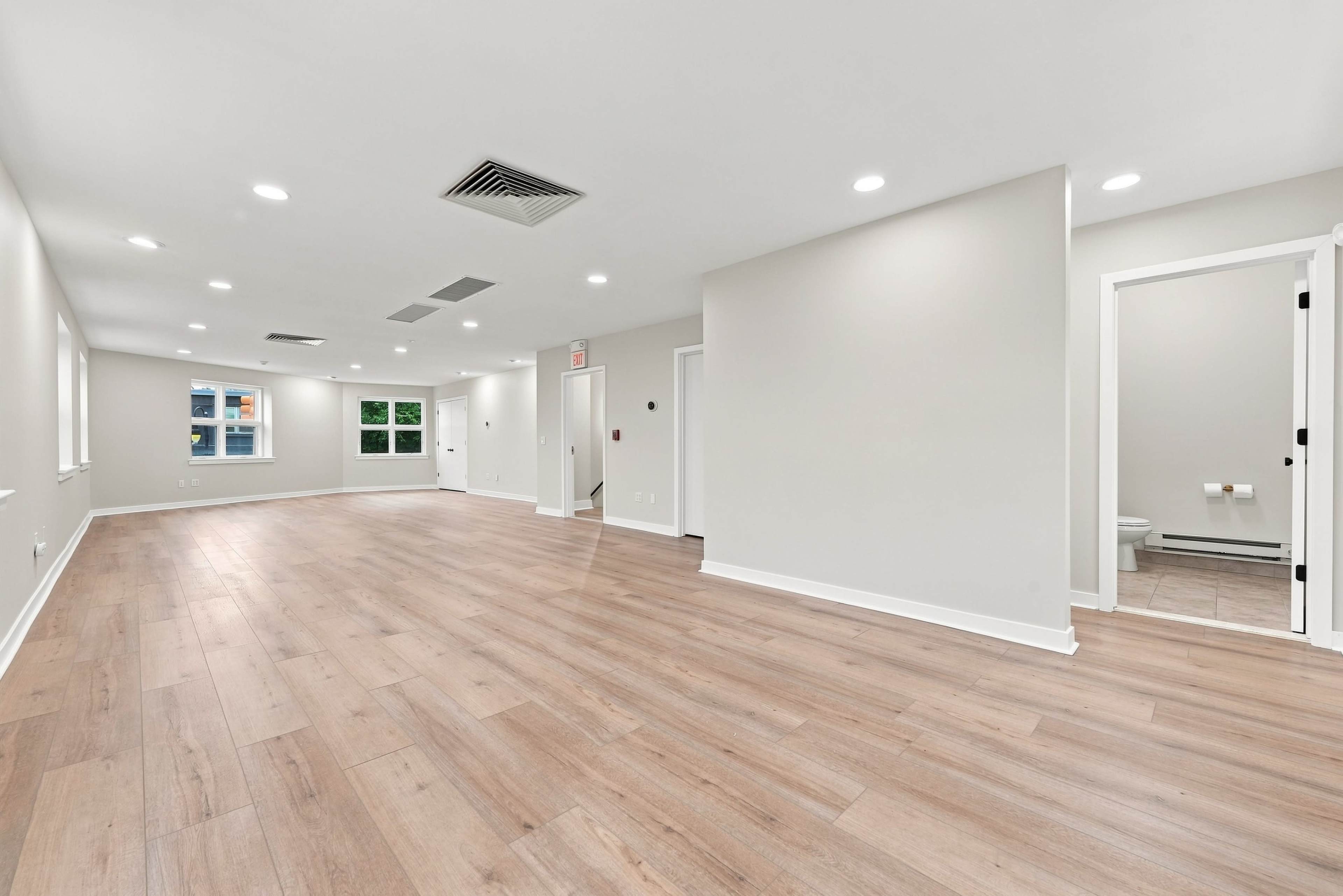 The image shows a spacious, empty room with light wood flooring and white walls, featuring multiple windows and a doorway leading to a bathroom.