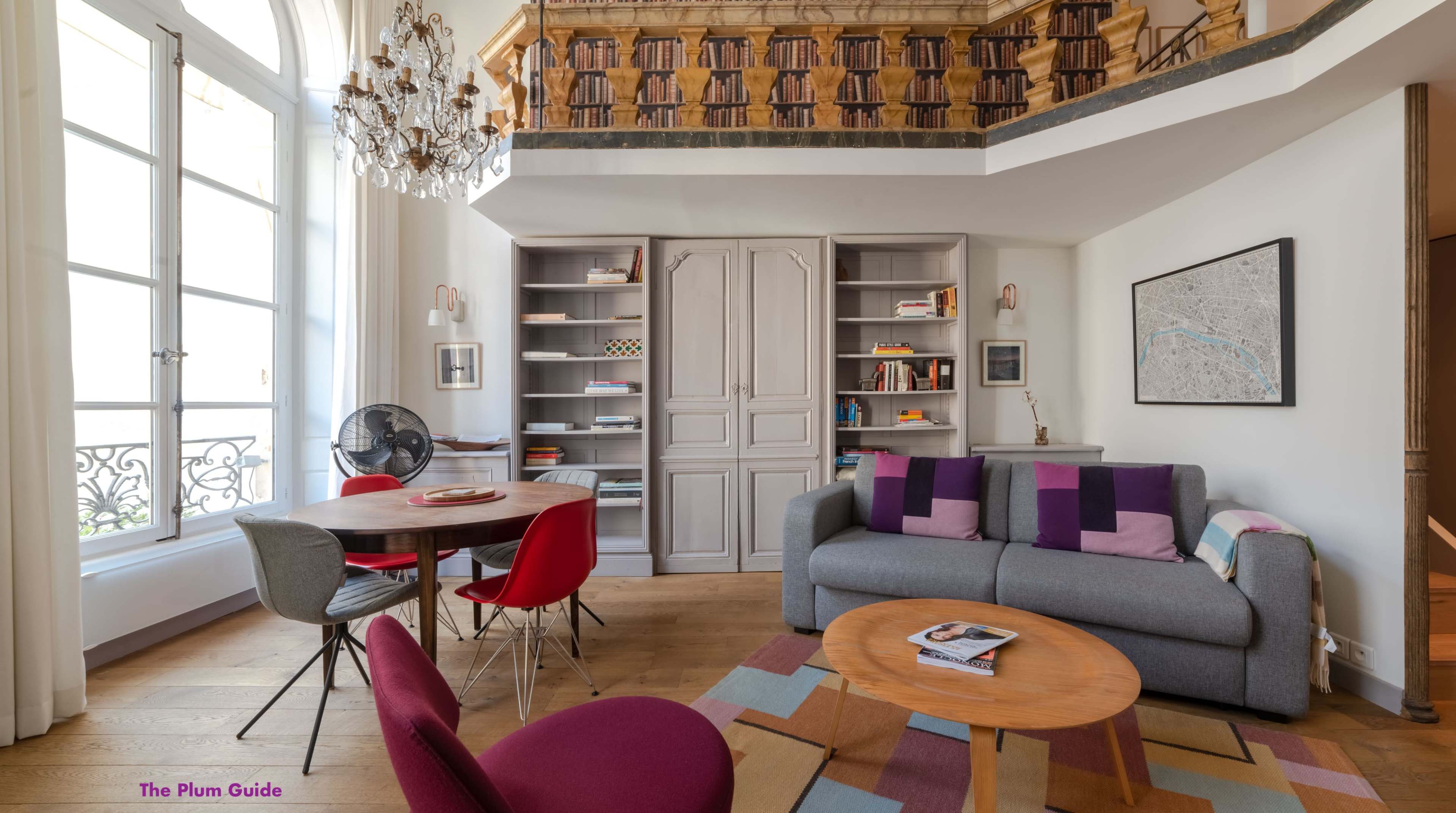 A cozy living room with a gray sofa, a circular wooden table surrounded by red and purple chairs, and a bookshelf on the wall.