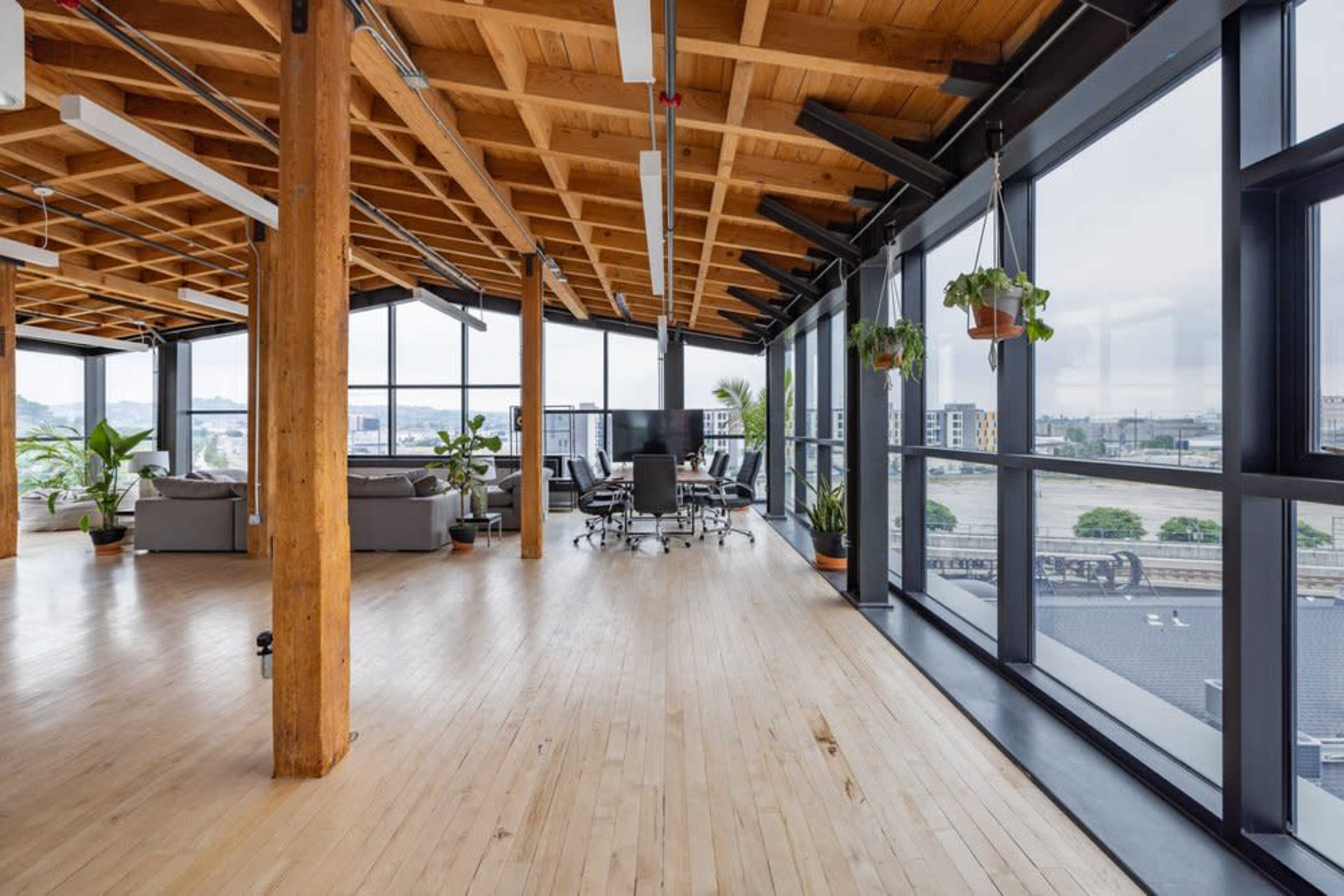 A spacious office interior features large windows, wooden beams, and a central meeting area surrounded by lush green plants.