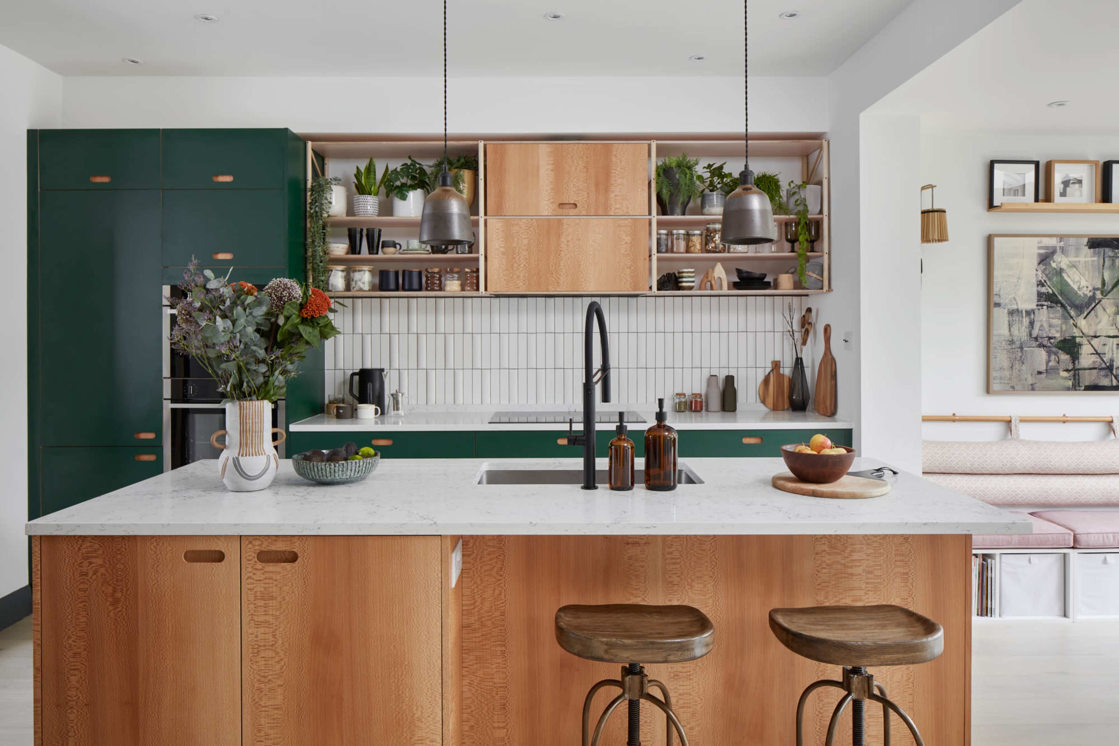 A modern kitchen features green cabinetry, wooden elements, a white countertop, and shelves adorned with plants and kitchenware.