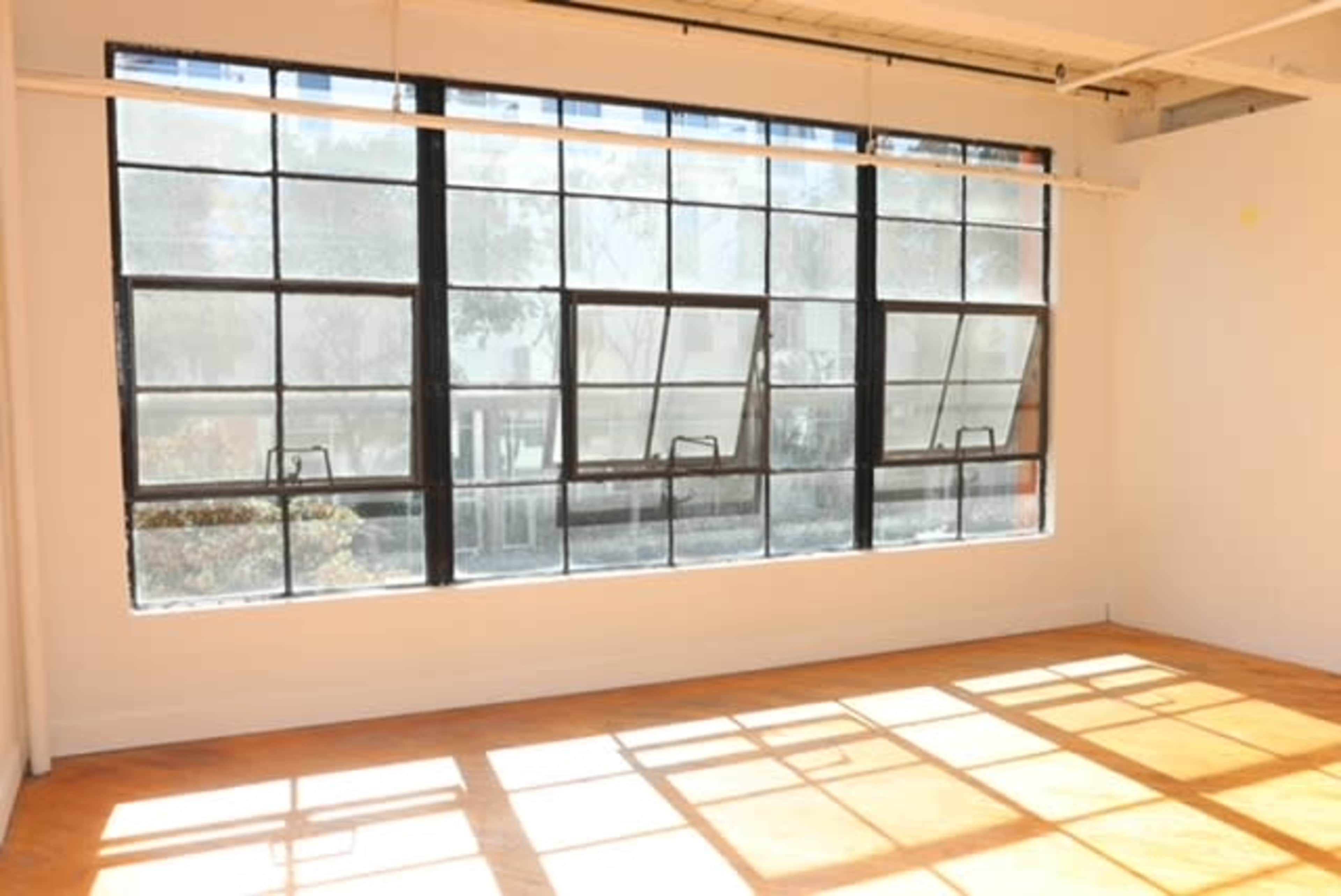 The image shows a bright room with large, open windows allowing sunlight to cast shadows on the wooden floor.