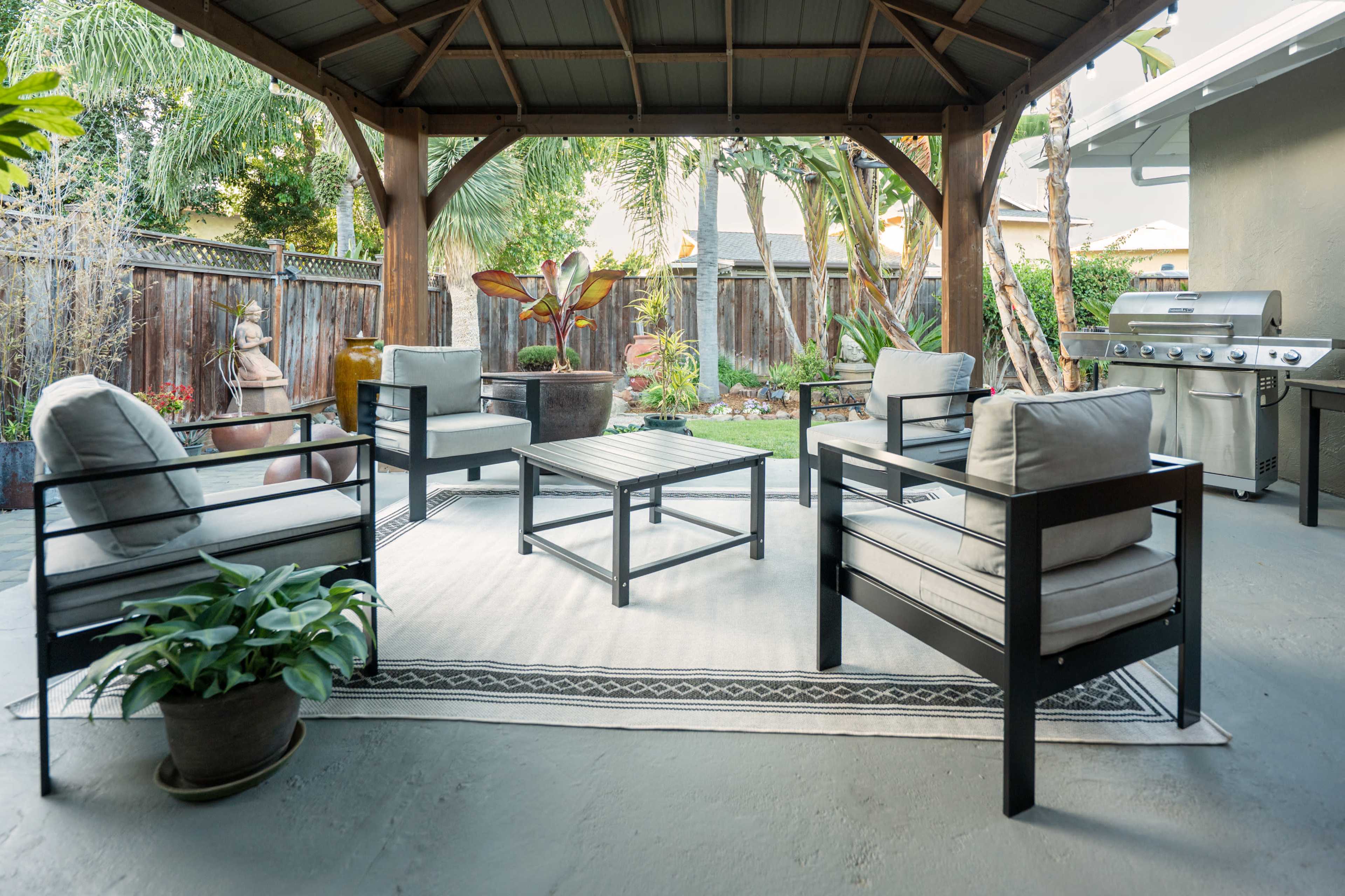 A shaded outdoor patio features four chairs surrounding a table, with a barbecue grill and greenery in the background.