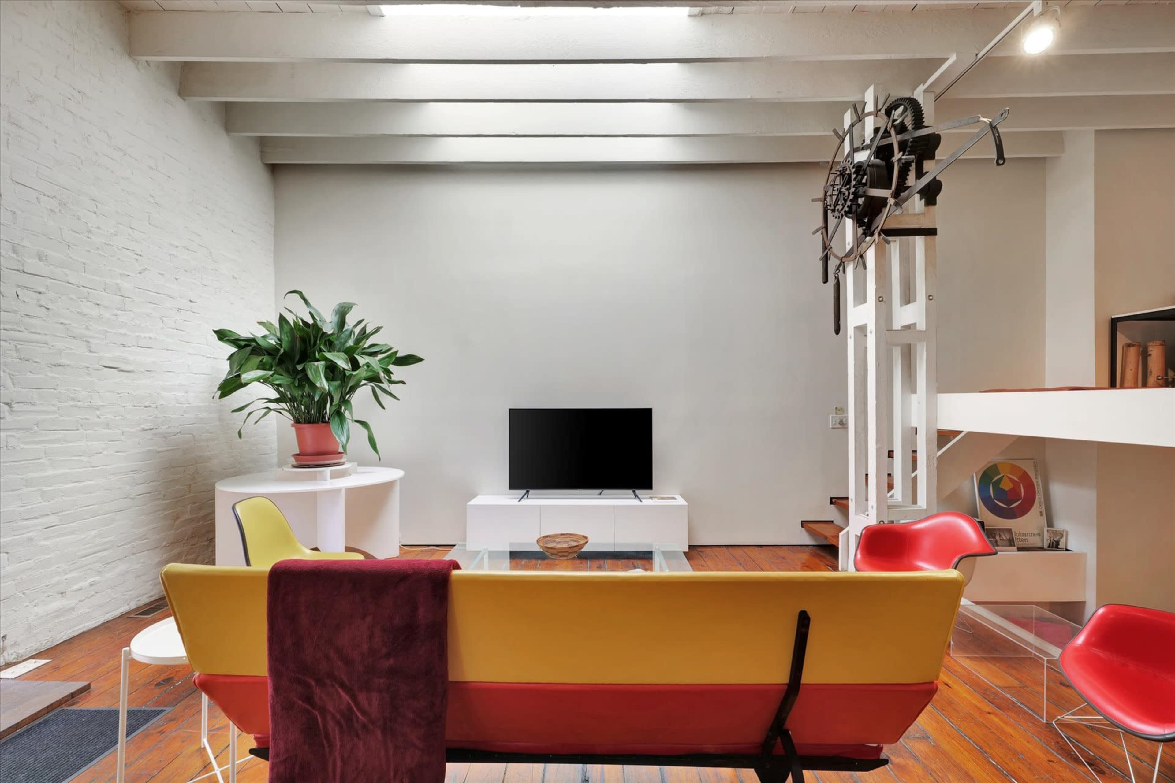 The image shows a modern living room with a yellow and black sofa, a glass coffee table, a television, and a large houseplant, all set against a backdrop of white brick walls and wooden flooring.