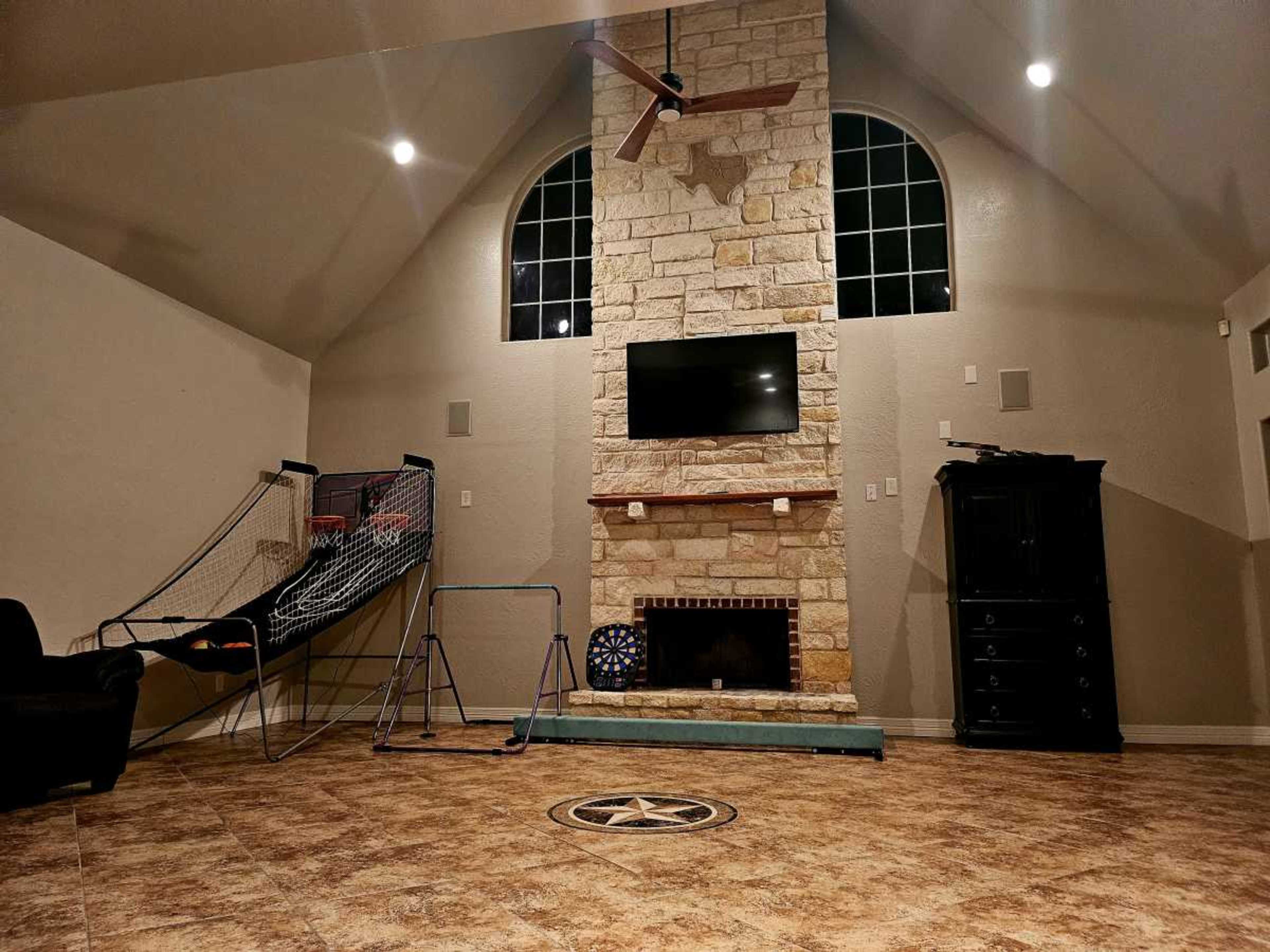 The image shows a spacious living area with a stone fireplace, a mounted TV, a wall-mounted basketball hoop, and a large window above the fireplace.