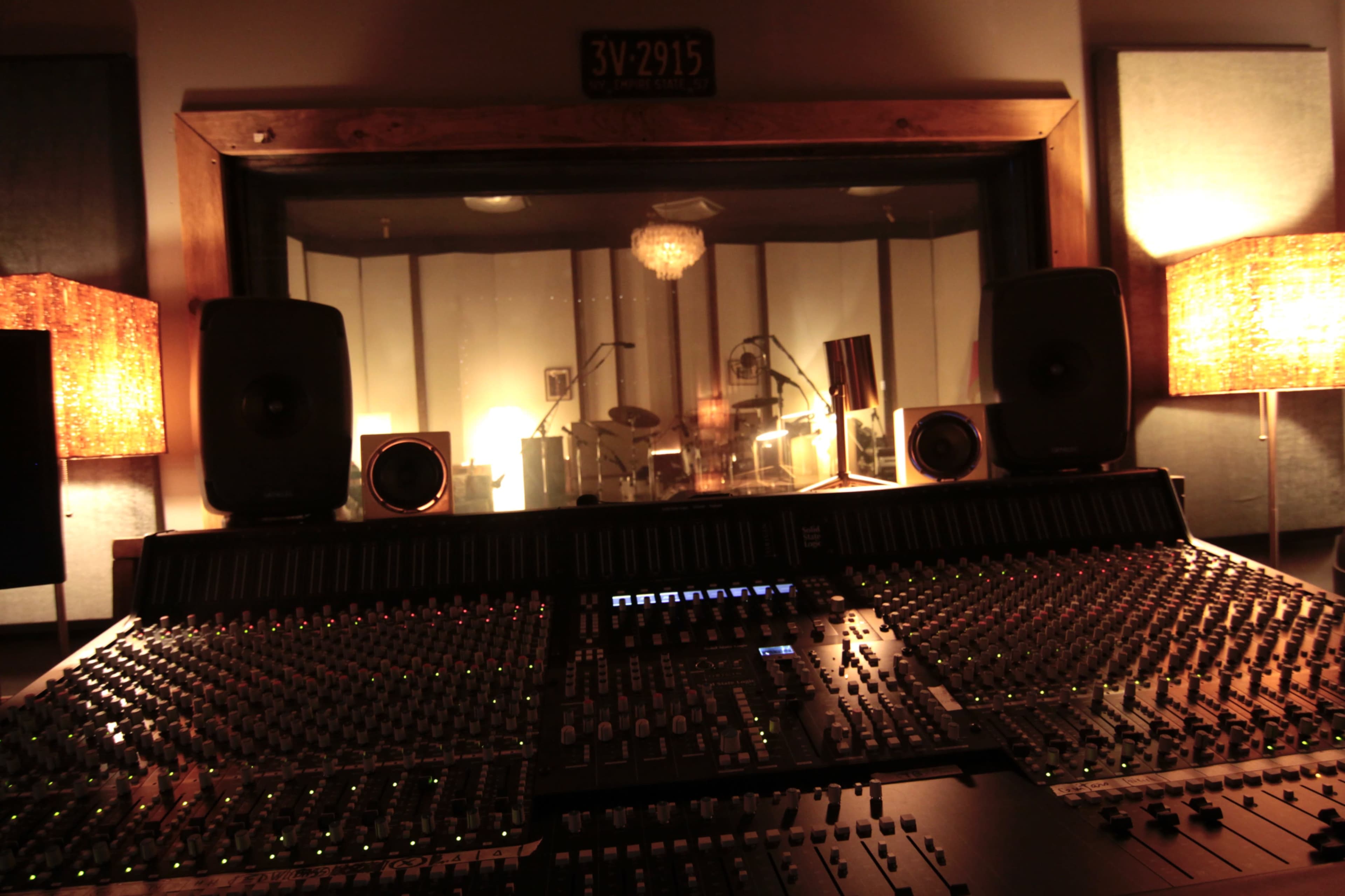 A recording studio control room is shown with a large mixing console in the foreground and a glass partition overlooking a soundproof room with instruments and lights.
