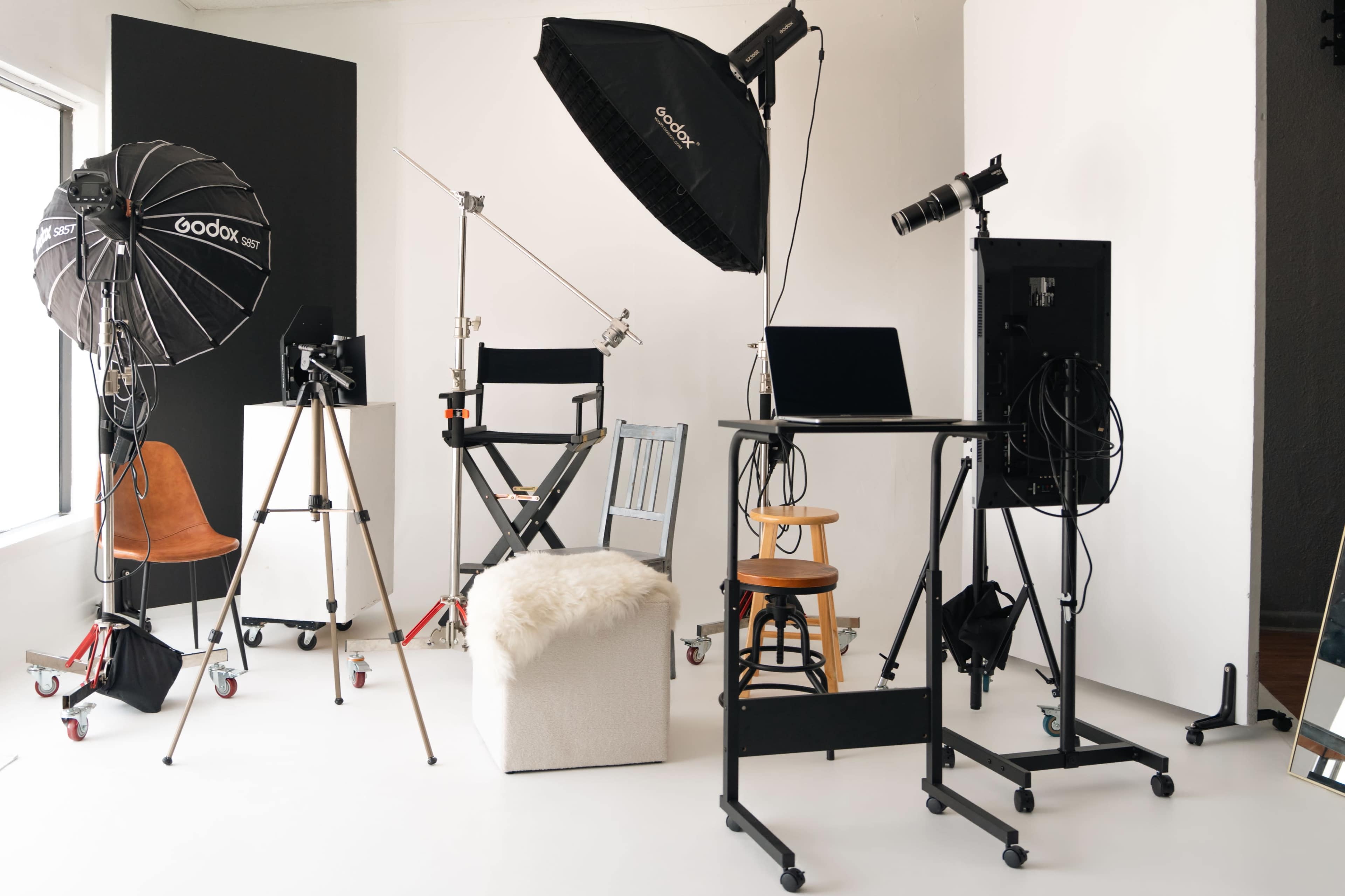 The image shows a photography studio setup with various lighting equipment, chairs, a computer, and a backdrop.