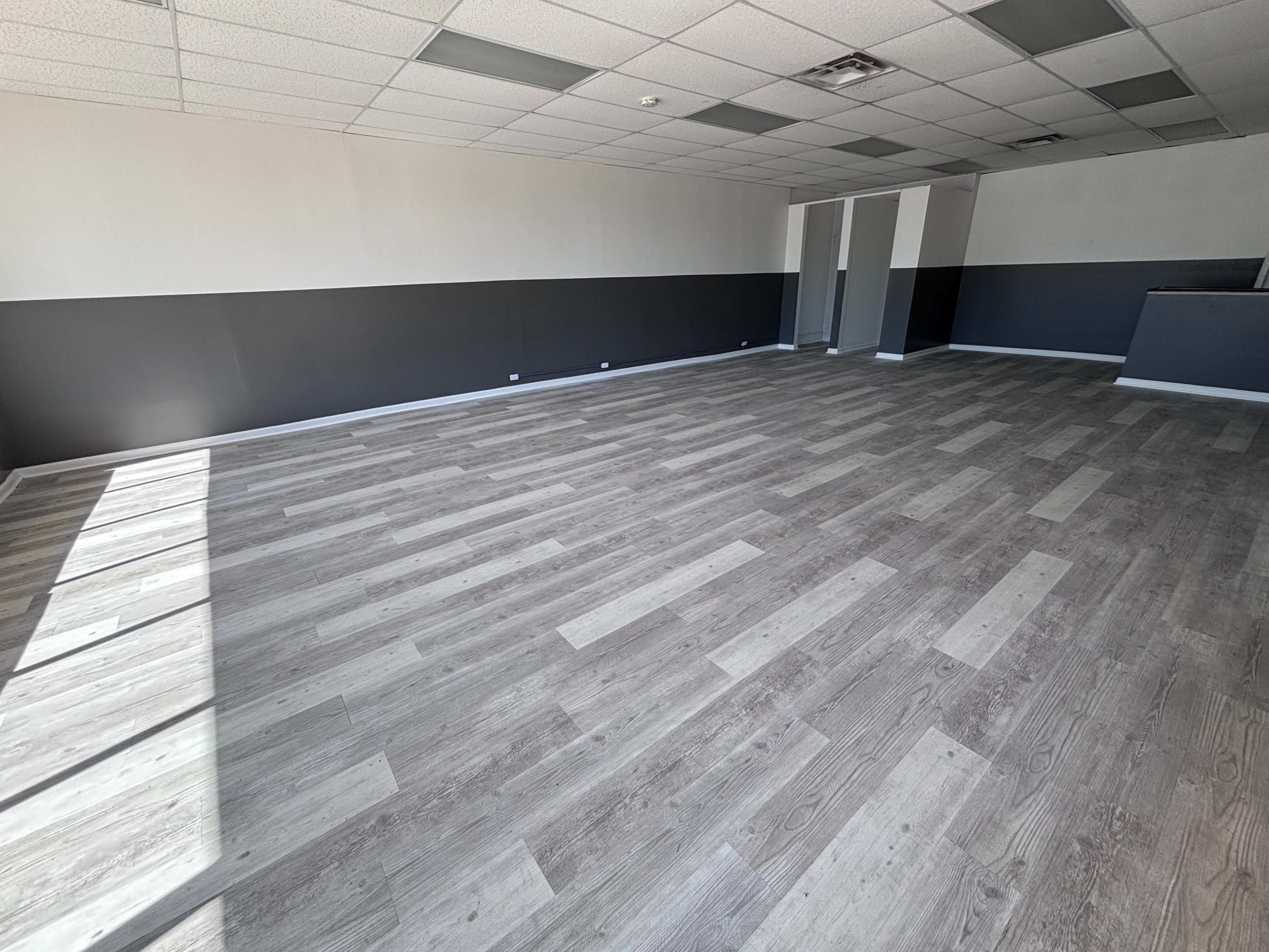 The image shows a vacant interior space with gray wooden flooring and light-colored walls featuring a horizontal black stripe.