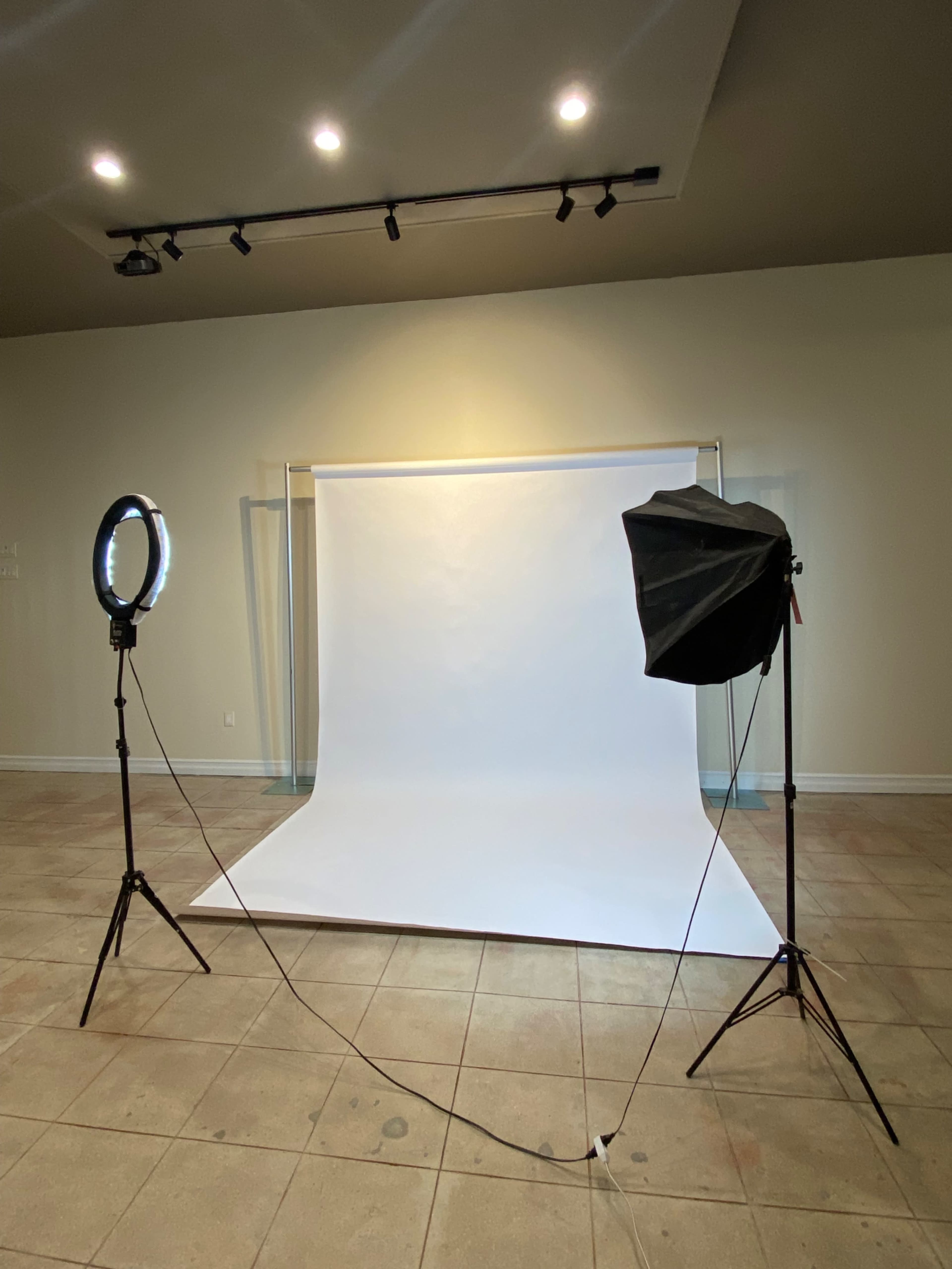 The image shows a photography setup with a white backdrop, a softbox light, and a ring light positioned in a spacious room with tiled flooring.