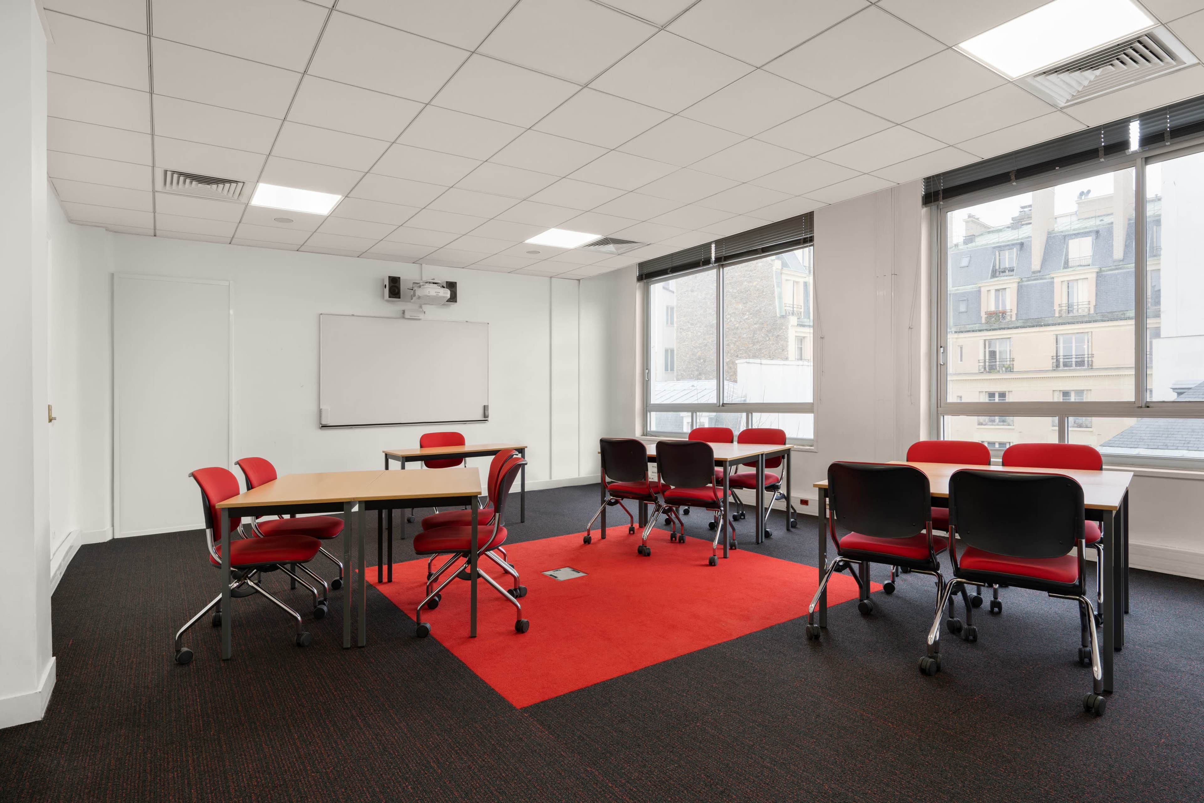 A modern meeting room with red chairs around several tables and large windows overlooking a cityscape.