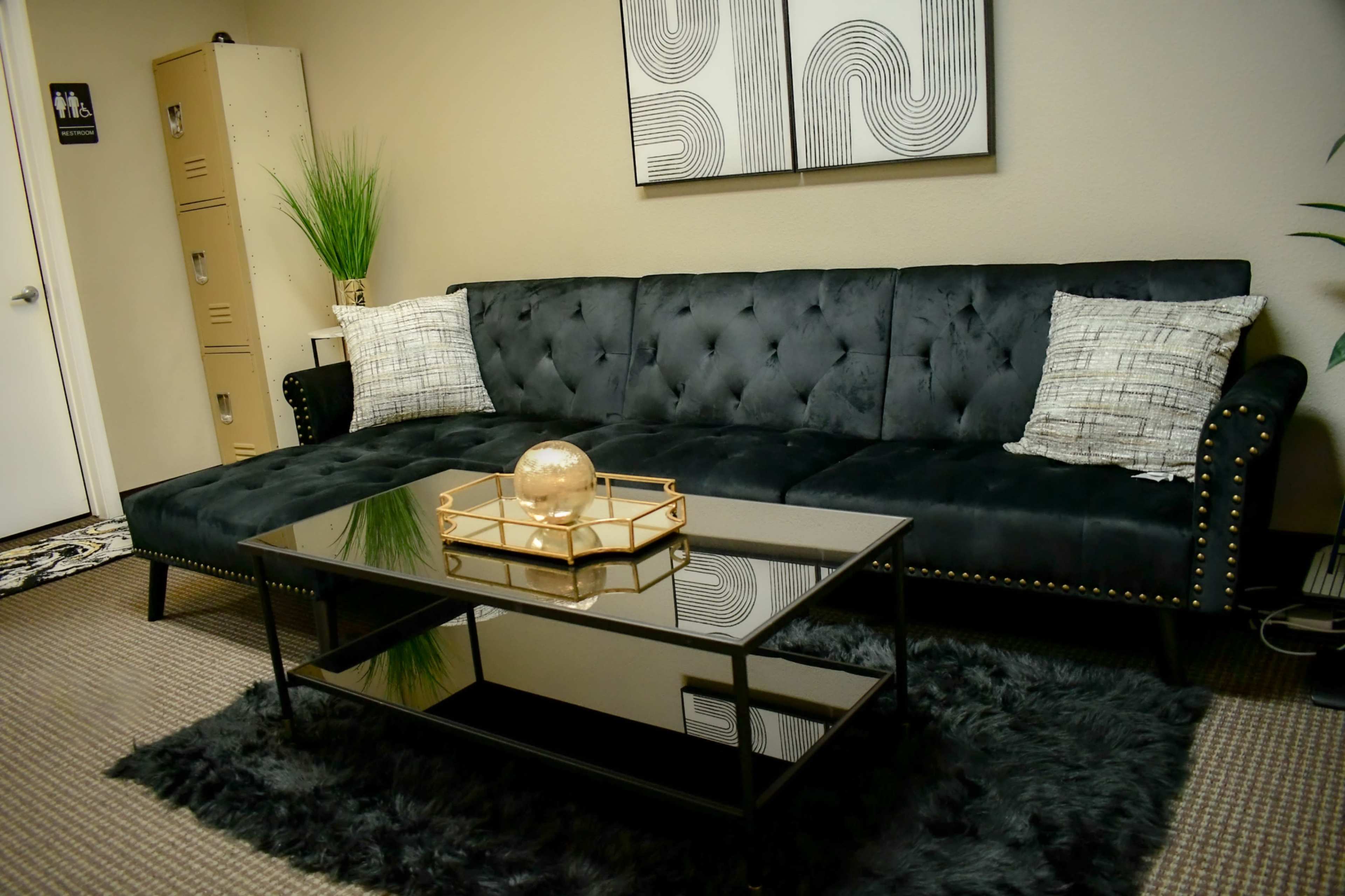 A modern living room with a black tufted sofa, a glass coffee table, and decorative elements like a plant and wall art.