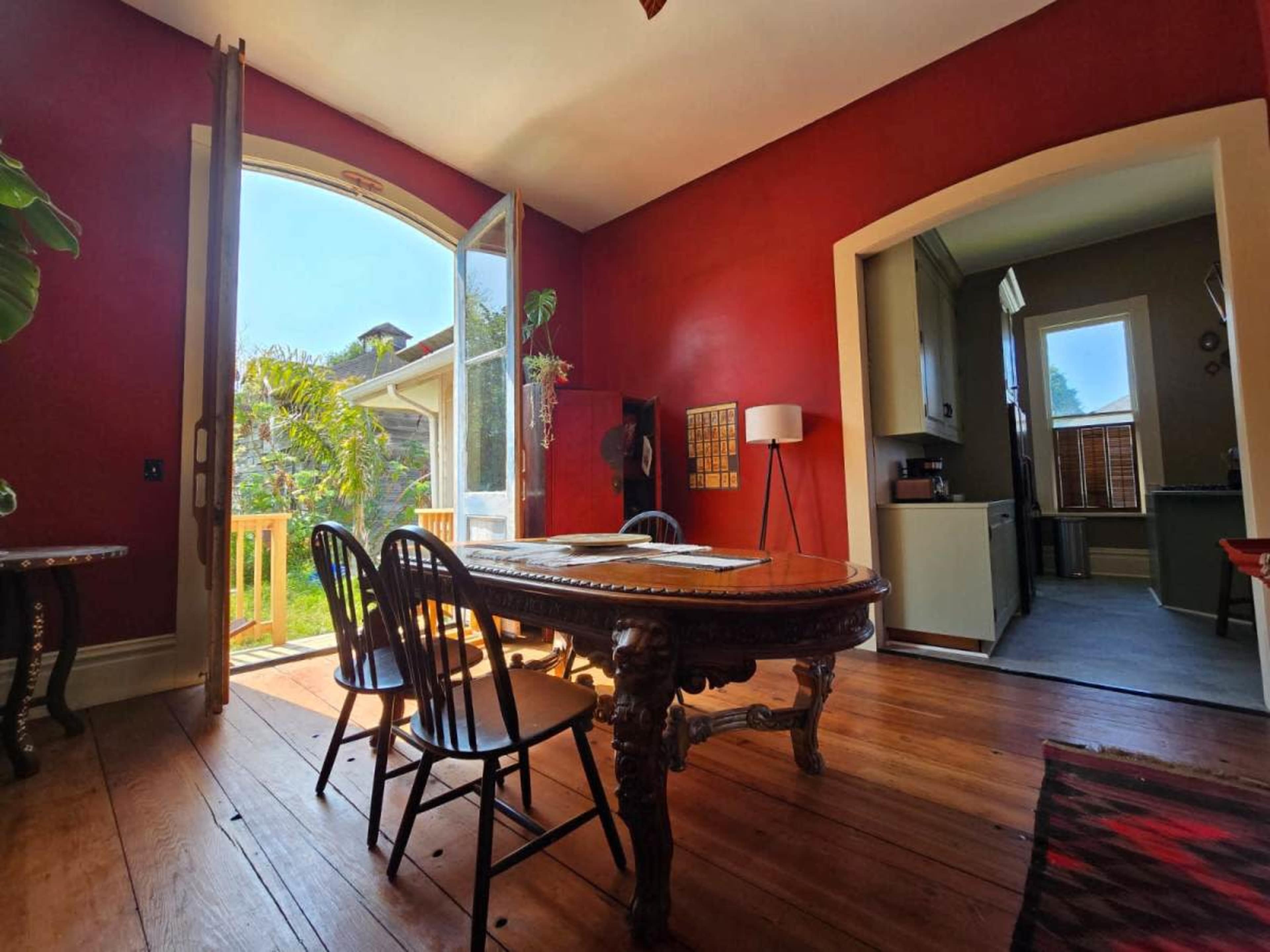 A dining area features a round table with three chairs, set against a red wall, and opens to a sunny balcony through large French doors.