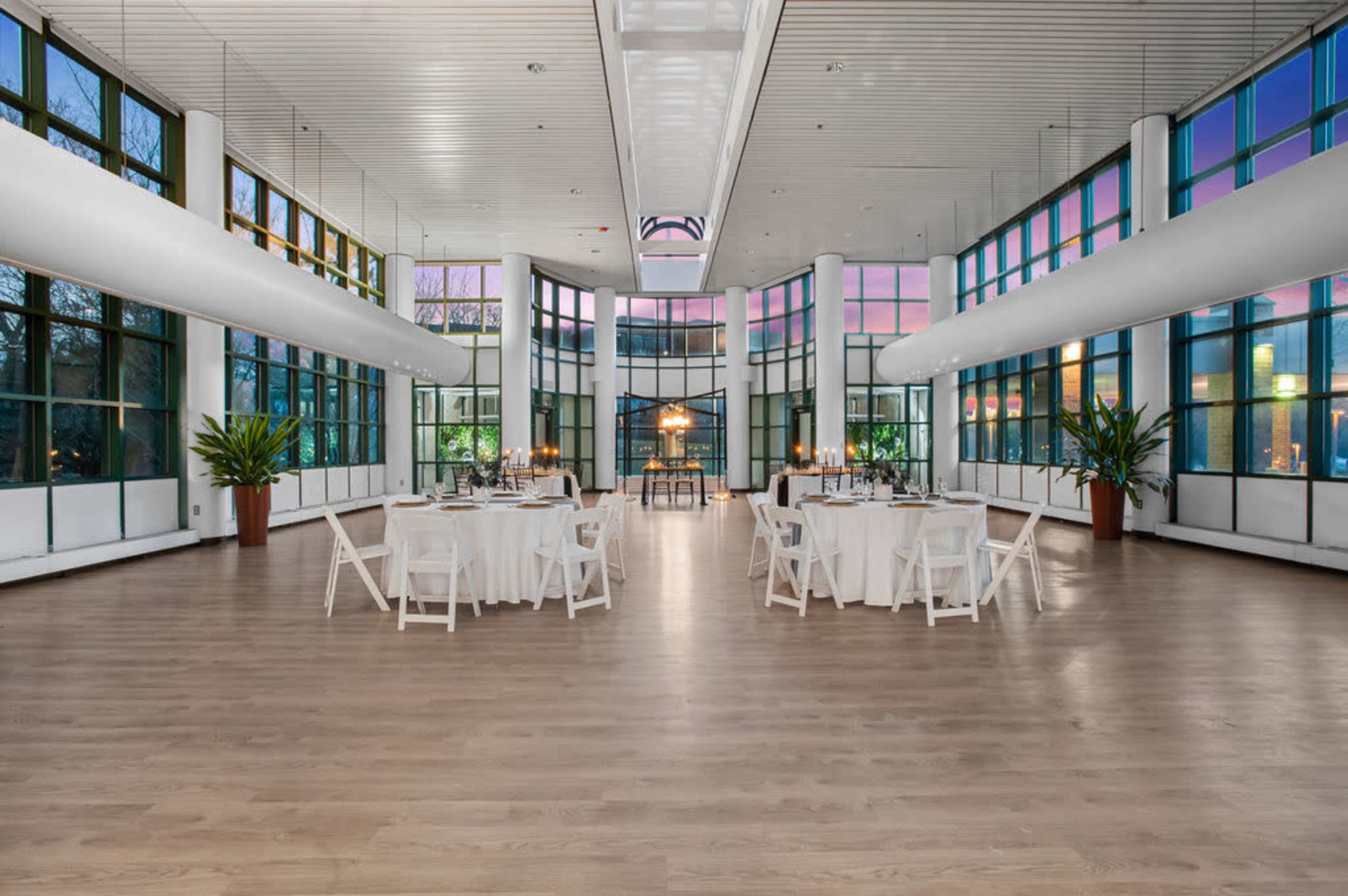 A spacious event room with large windows, featuring white tables set for a gathering and potted plants in the corners.