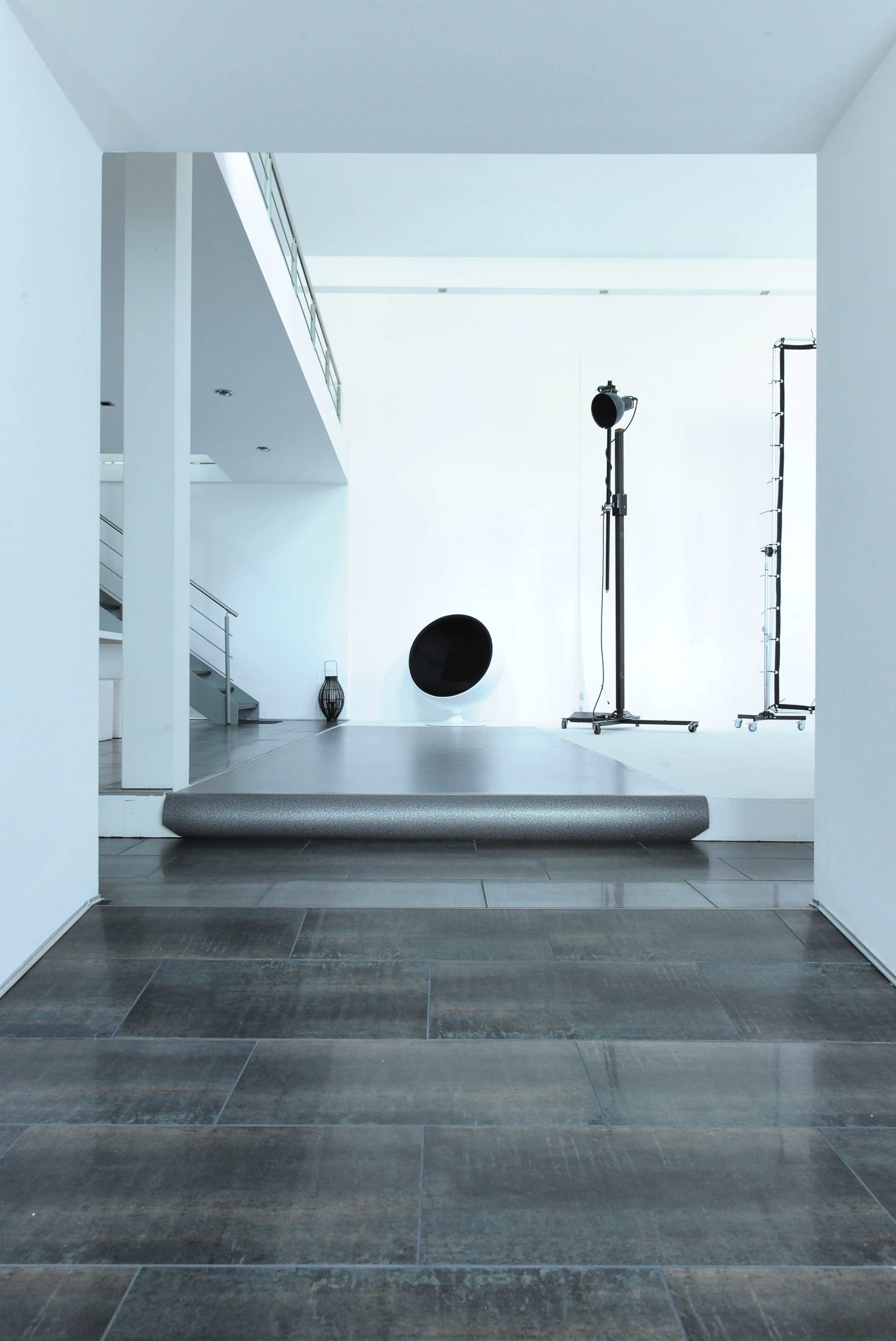 The image depicts a modern indoor space featuring a smooth, protruding platform leading towards a minimalist setup with a circular wall opening and a standing light fixture.
