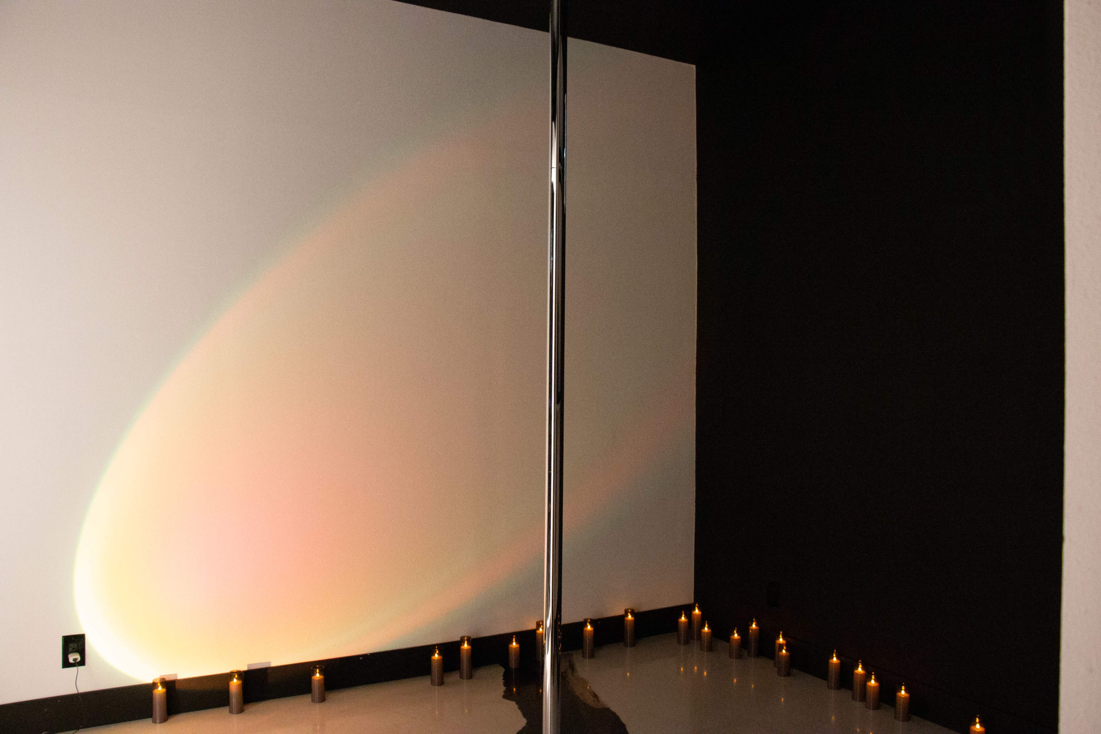 A polished dance pole stands between a white wall with a gradient of light and a black wall, surrounded by a circle of flickering candles on the floor.