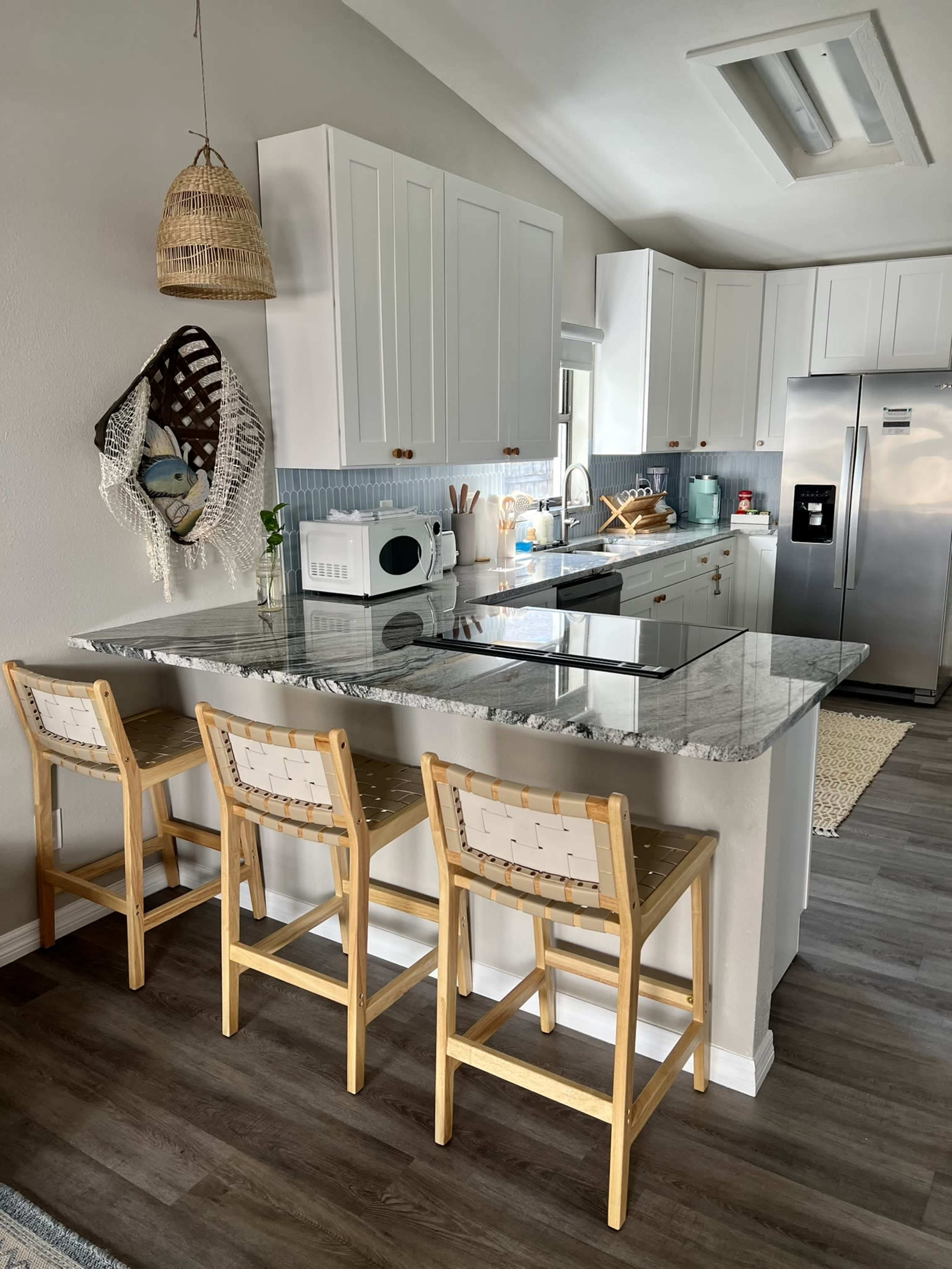 A modern kitchen features white cabinetry, a granite countertop island with three wooden stools, and stainless steel appliances, all enhanced by natural light.
