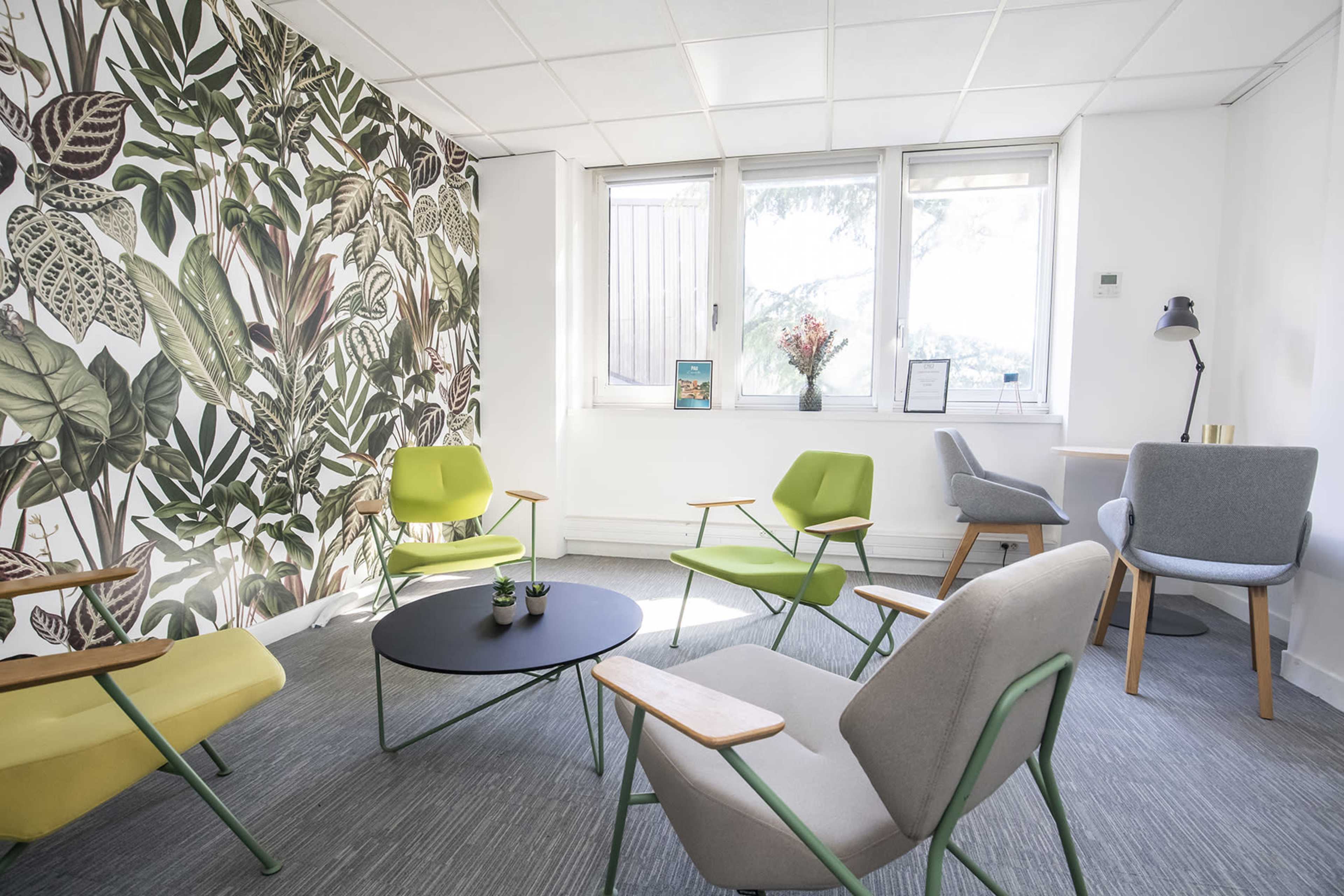 A modern seating area features green and gray chairs arranged around a black coffee table, with a plant-themed wallpaper and large windows letting in natural light.