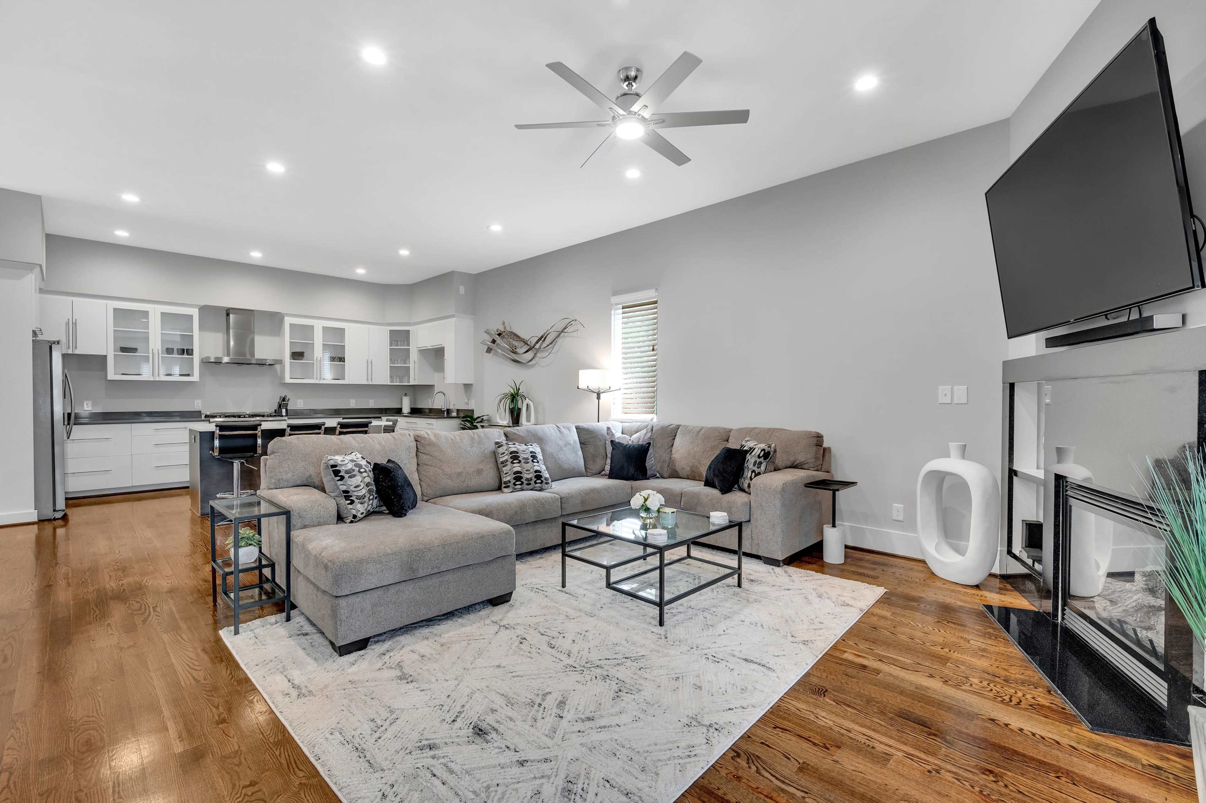 A spacious living room features a large gray sectional sofa, a coffee table, and a flat-screen TV mounted on the wall, with a kitchen visible in the background.