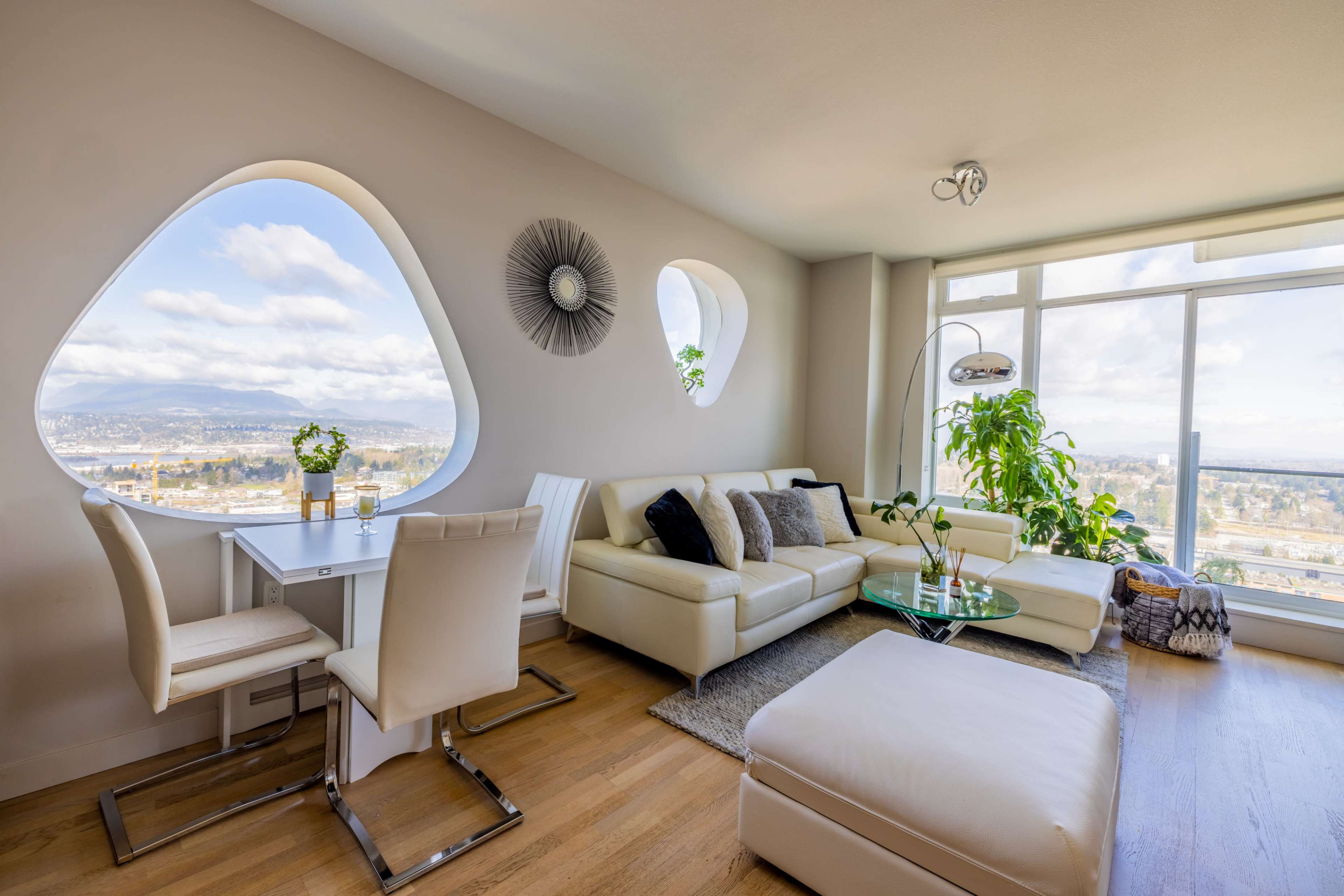 A modern living room features a white leather sofa, a glass coffee table, and a dining area with a view through unique, rounded windows.