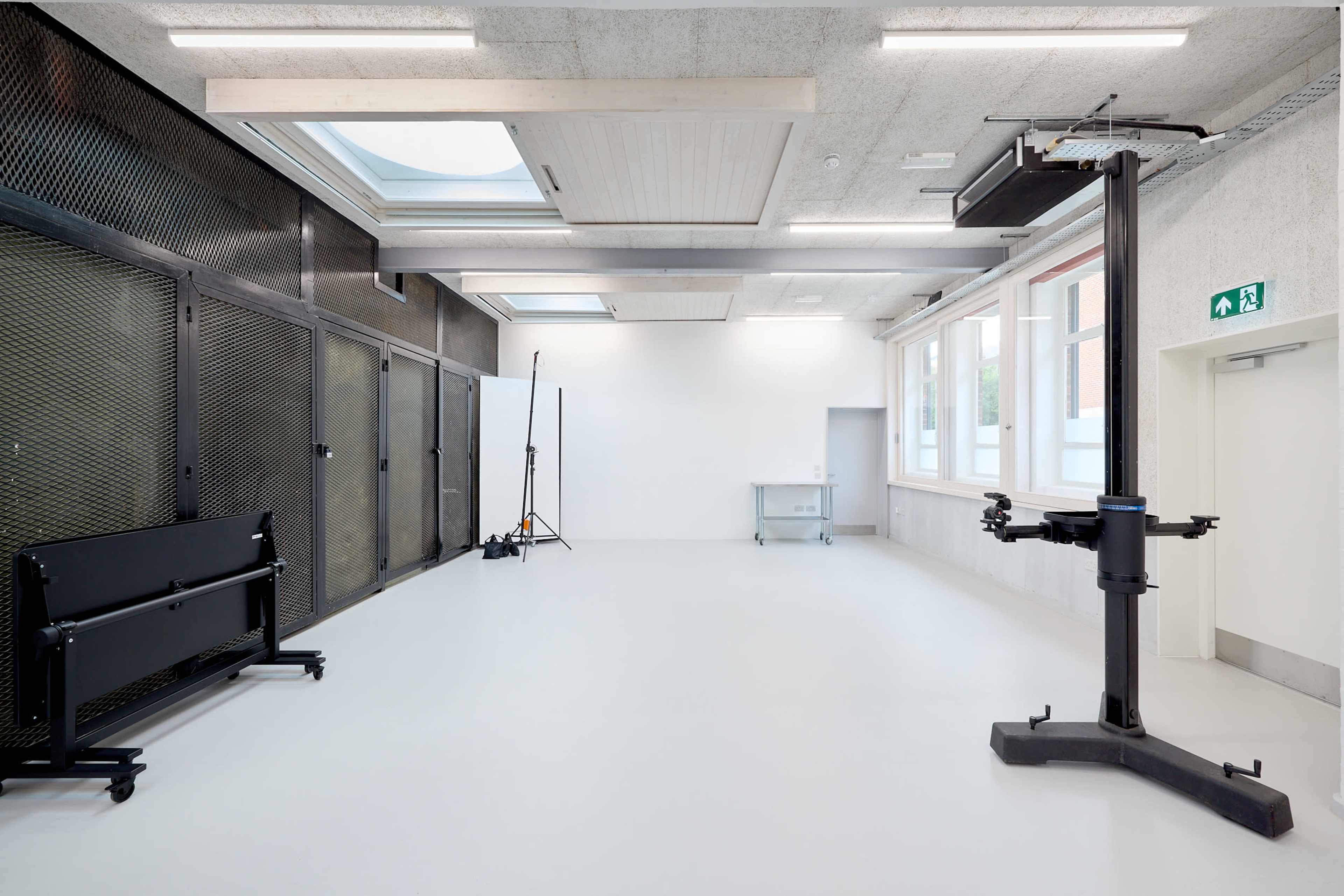 The image shows a spacious, well-lit room featuring a high ceiling, a large overhead skylight, and a black lift apparatus along with various photography equipment and a rolling cart.