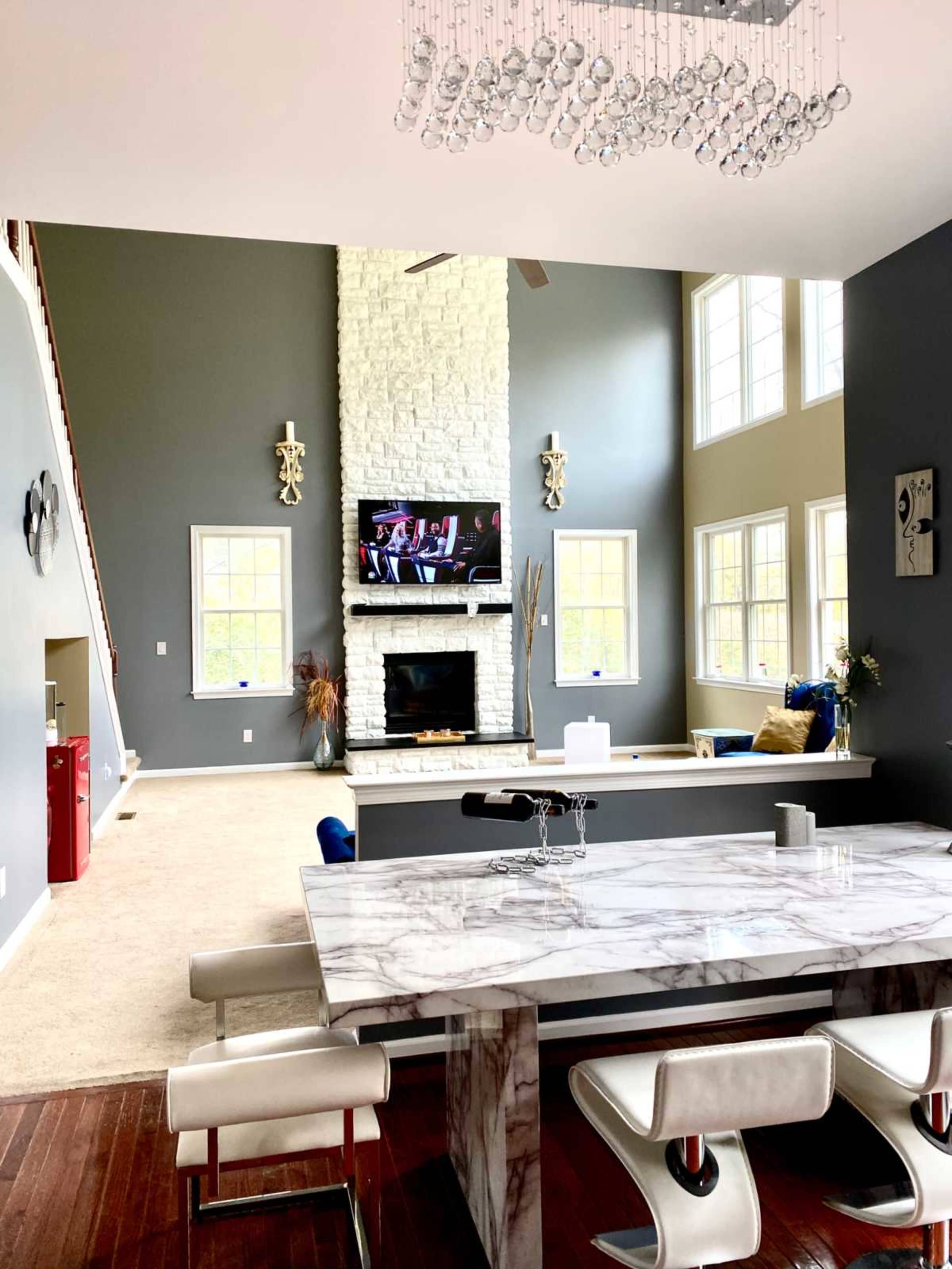The image features a modern living space with a high ceiling, a marble dining table, a stone fireplace, and large windows.