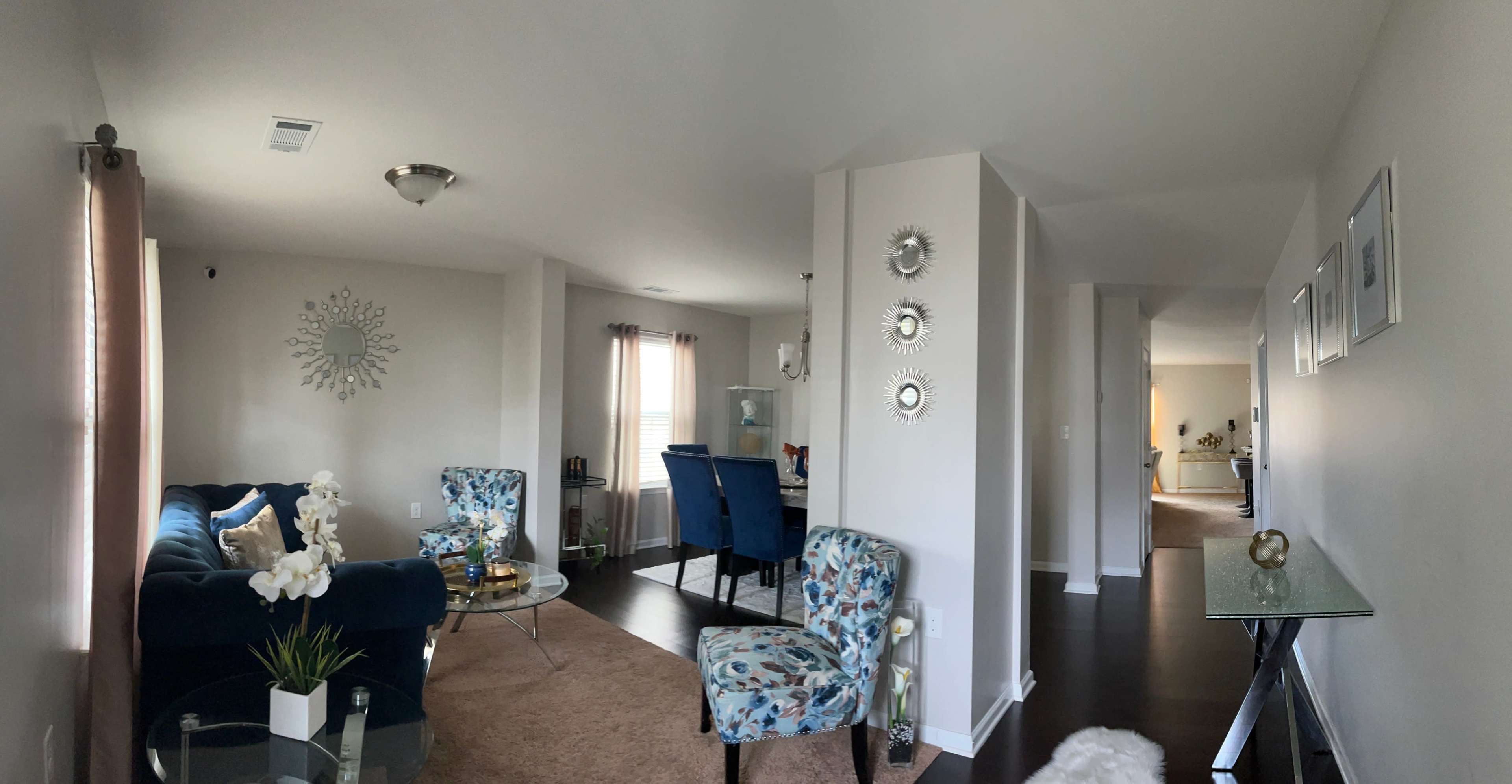A modern living room with floral-patterned chairs, a glass coffee table, and a clear view into an adjacent dining area and hallway.
