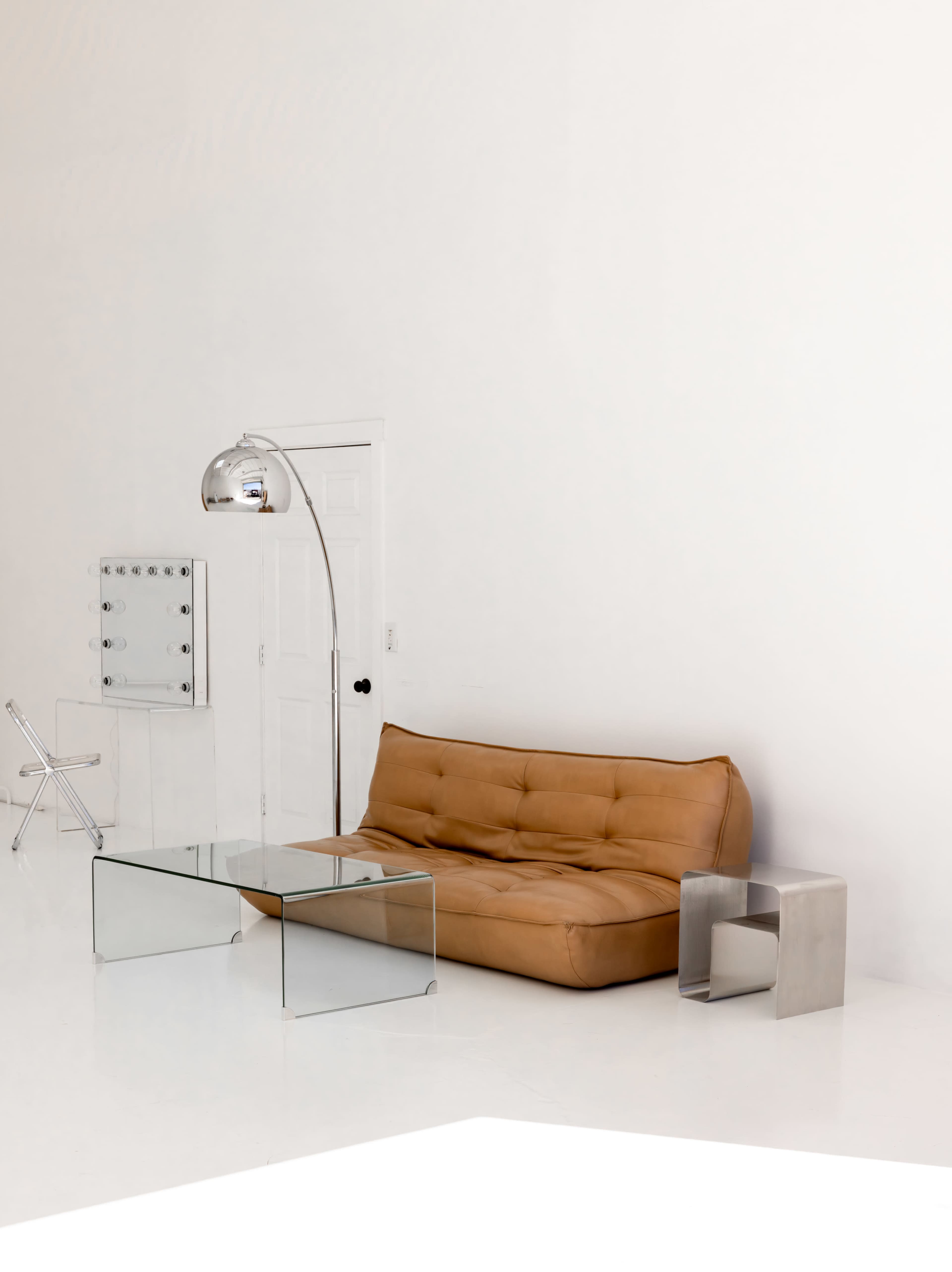 The scene features a modern, minimalist living room with a brown sofa, a glass coffee table, a metallic side table, and a silver arc floor lamp against a white backdrop.