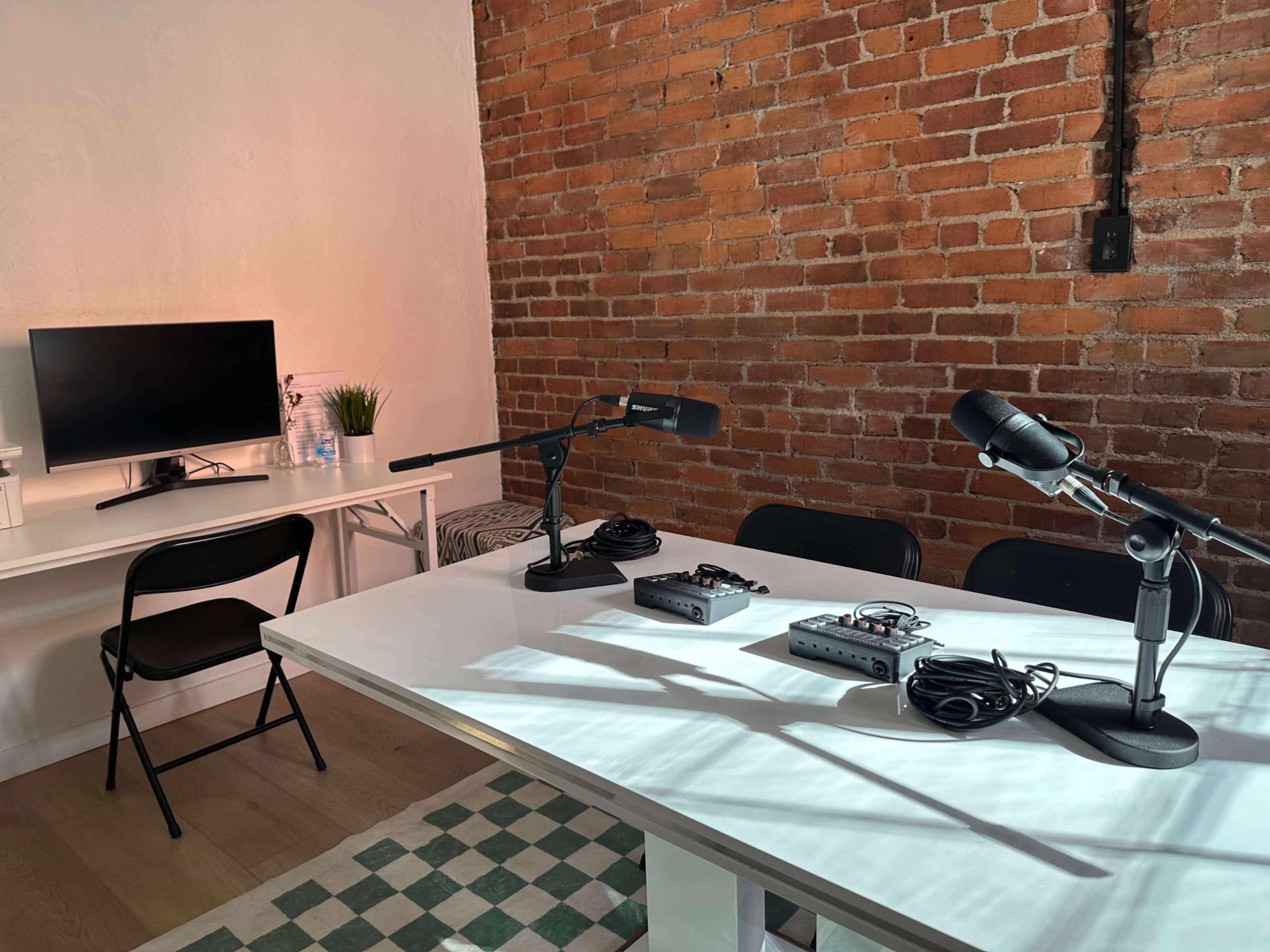A bright room features a white table with two microphones, audio equipment, a monitor, and a black chair, against a backdrop of exposed brick walls.