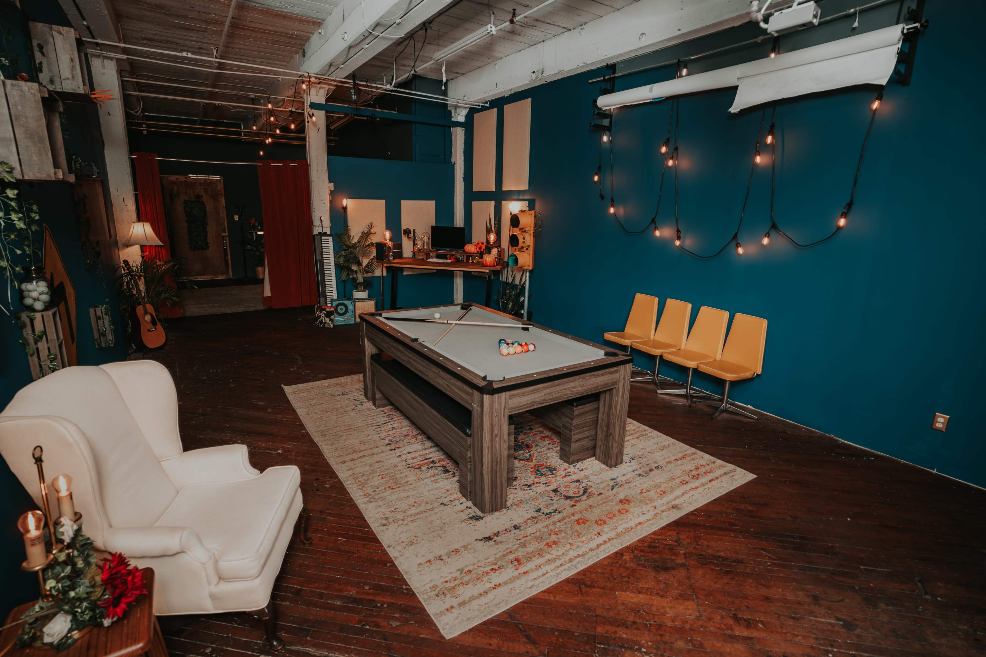 A game room featuring a pool table in the center, surrounded by yellow chairs, a cozy armchair, and decorated with string lights on the walls.