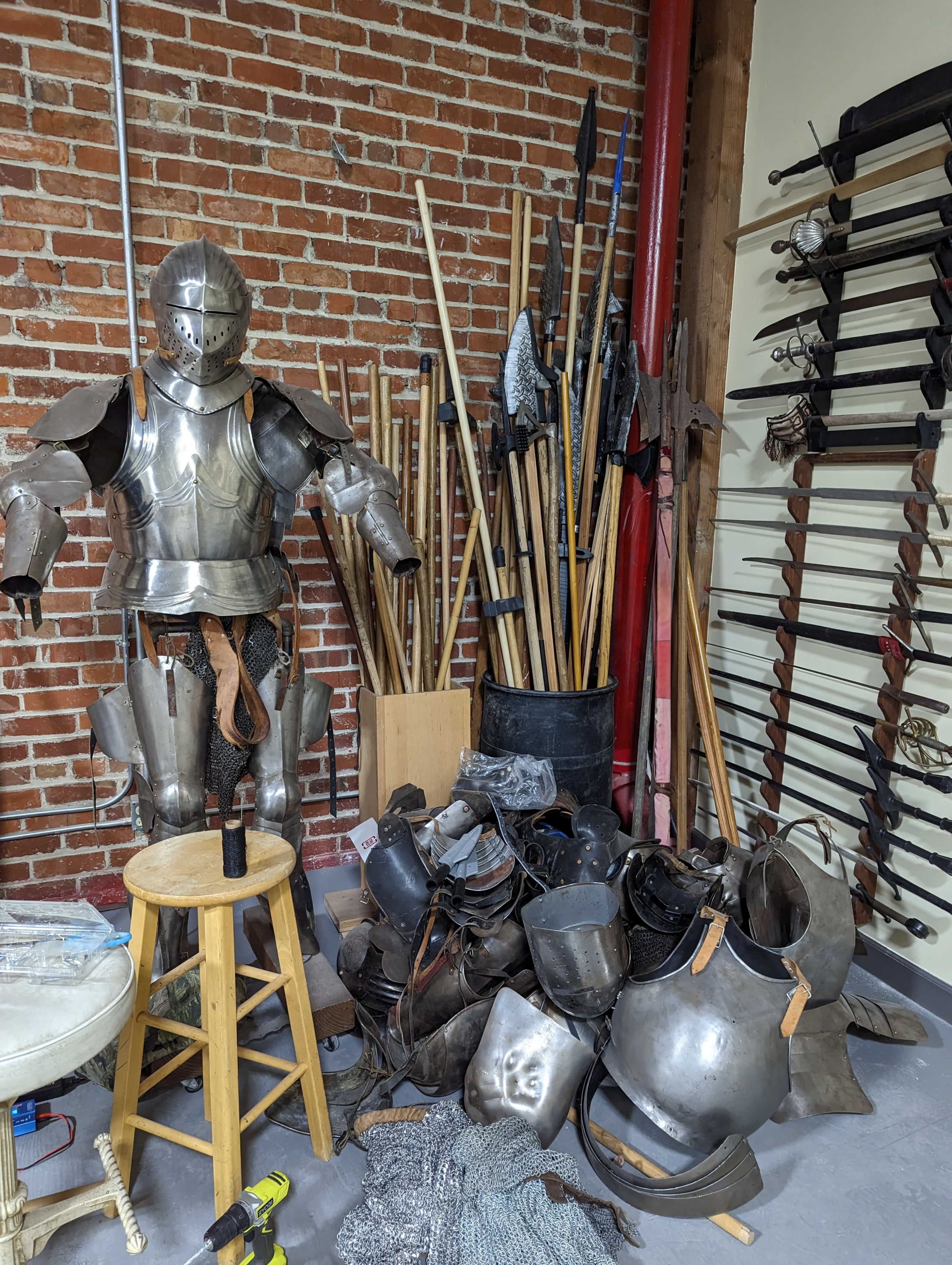 A suit of medieval armor stands on a stool next to a pile of various metallic armor pieces and wooden spears against a brick wall.