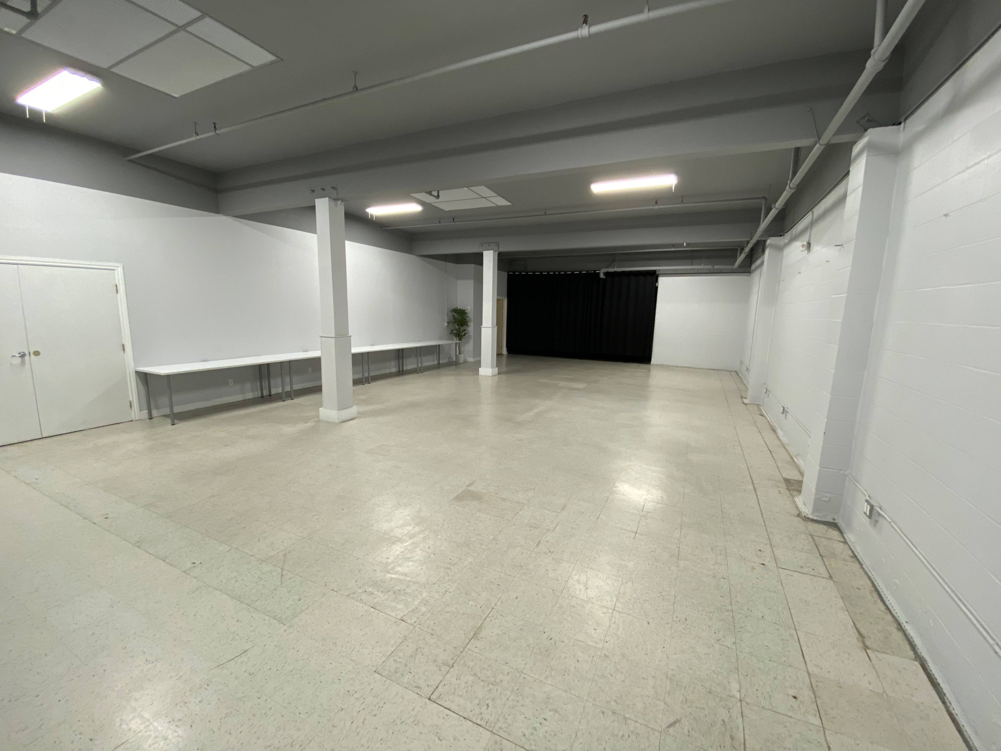 An empty room with gray walls, a tiled floor, and two long tables along one side, leading to a black curtain at the far end.