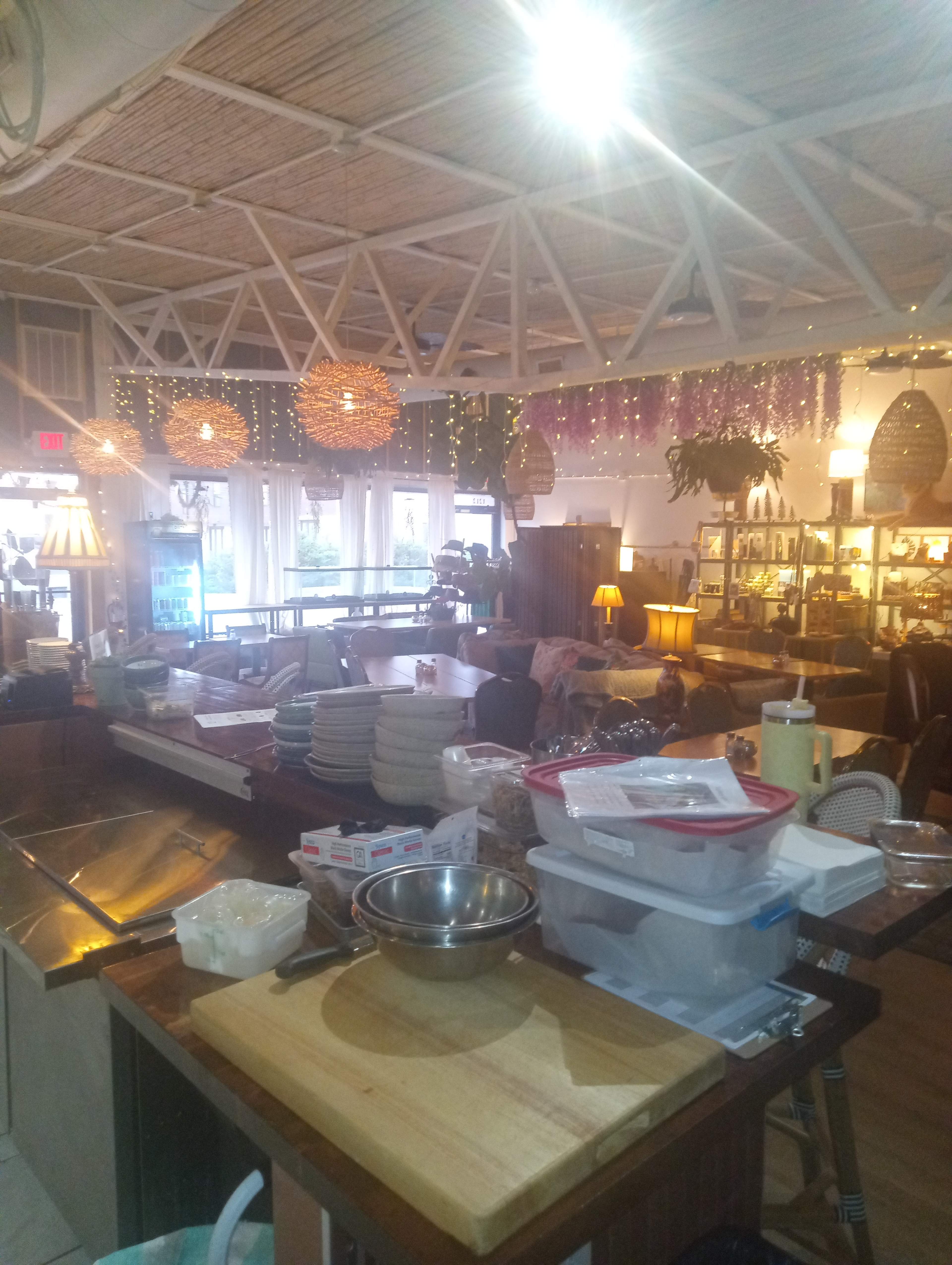 The image shows a cozy restaurant interior with a combination of kitchen equipment, tables, decorative lights, and various dishware arranged on surfaces.